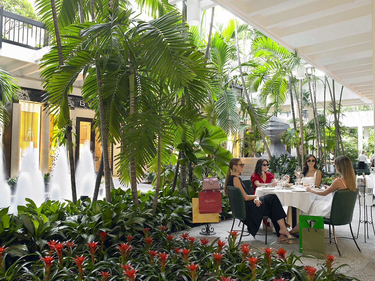 This image showcases a stylish outdoor dining area located on a patio or balcony. The scene is lush with tropical plants, including palm trees and red bromeliads, creating a relaxed and luxurious atmosphere. The presence of high-end retail store frontage suggests the property is situated in a upscale shopping district or possibly part of a mixed-use development aimed at providing a sophisticated living experience.