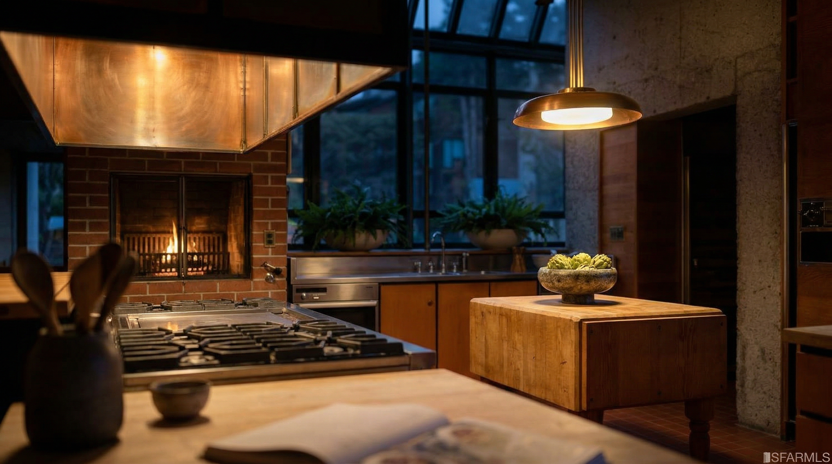 This sophisticated kitchen features a unique blend of industrial and rustic design elements, highlighted by a large copper hood and a cozy fireplace integrated into the brick wall. The space includes a substantial butcher-block island, warm wood cabinetry, and sleek stainless steel appliances, creating a welcoming yet high-end culinary environment. Large windows with an architectural grid overlook greenery, bathing the kitchen in natural light while a pendant light provides soft, ambient illumination.