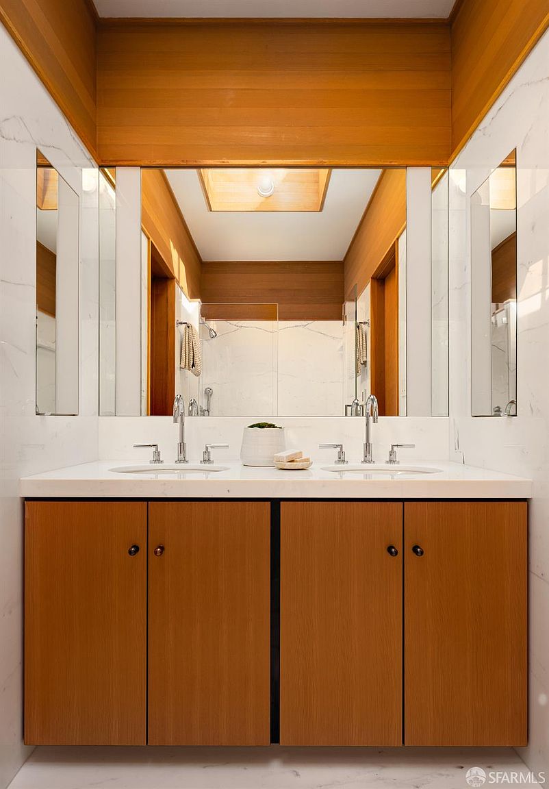 This is a well-lit primary bathroom featuring a double vanity with a white countertop and wooden cabinets. The space is adorned with large mirrors and marble-like wall tiles, creating a clean and modern aesthetic. A skylight provides natural light, enhancing the bright and airy feel of the bathroom.