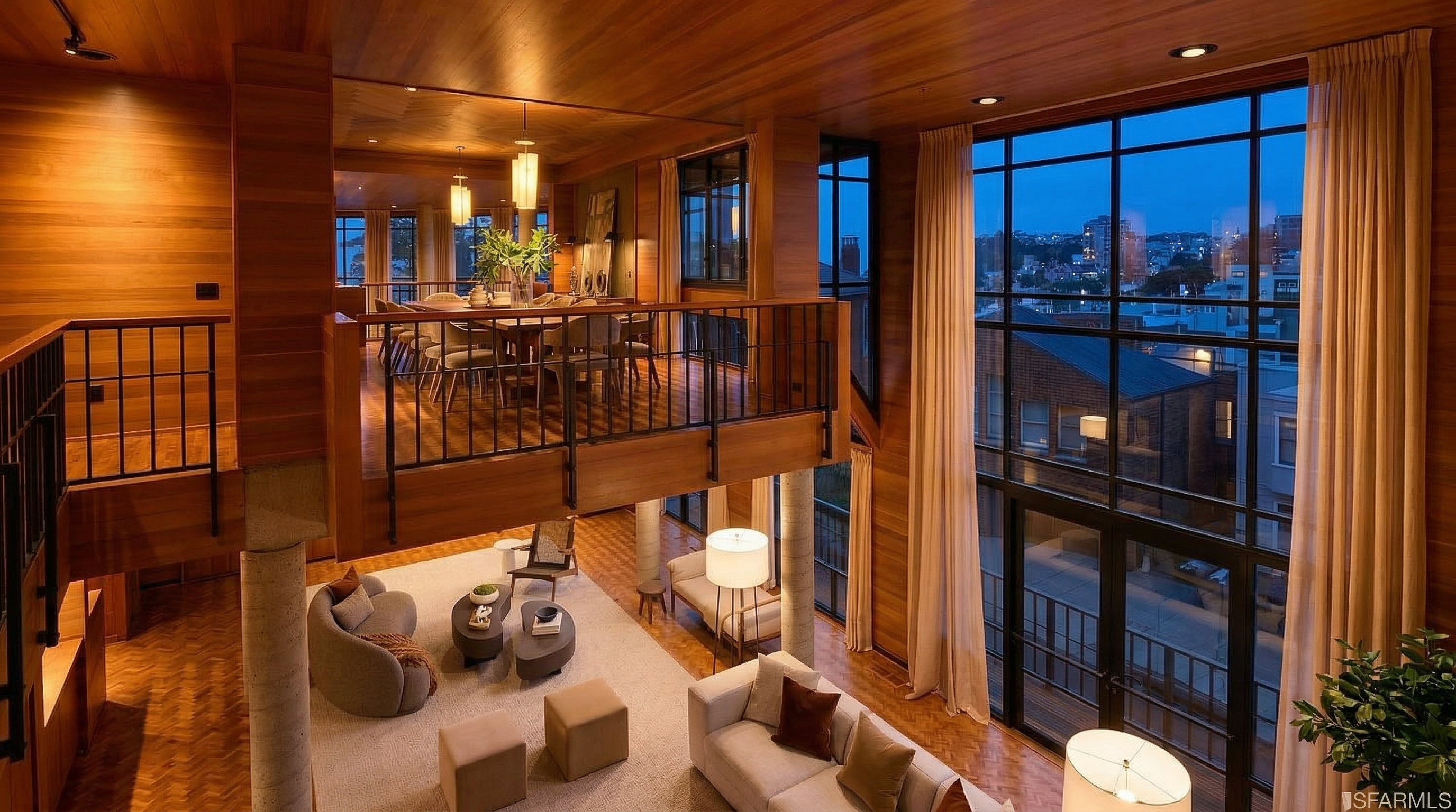 This is a high-angle shot of a luxurious, multi-level living room featuring warm wood paneling and large windows that offer a view of the cityscape. The space is furnished with modern sofas, chairs, and coffee tables arranged on a light-colored rug, creating a comfortable and inviting atmosphere. The open layout and natural light enhance the sense of spaciousness and elegance.