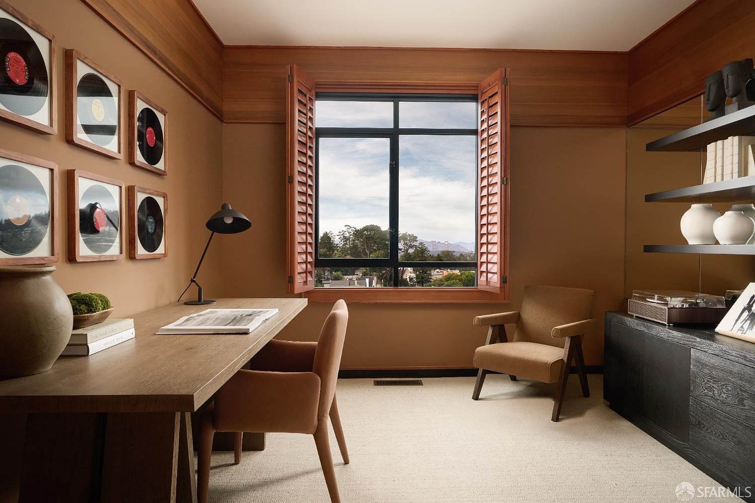 This interior shot showcases a well-appointed office or study. The room features a wooden desk with a black lamp, a comfortable armchair, and built-in shelving. The walls are painted in a warm brown tone, complemented by wooden trim and window shutters, creating a cozy and sophisticated workspace.