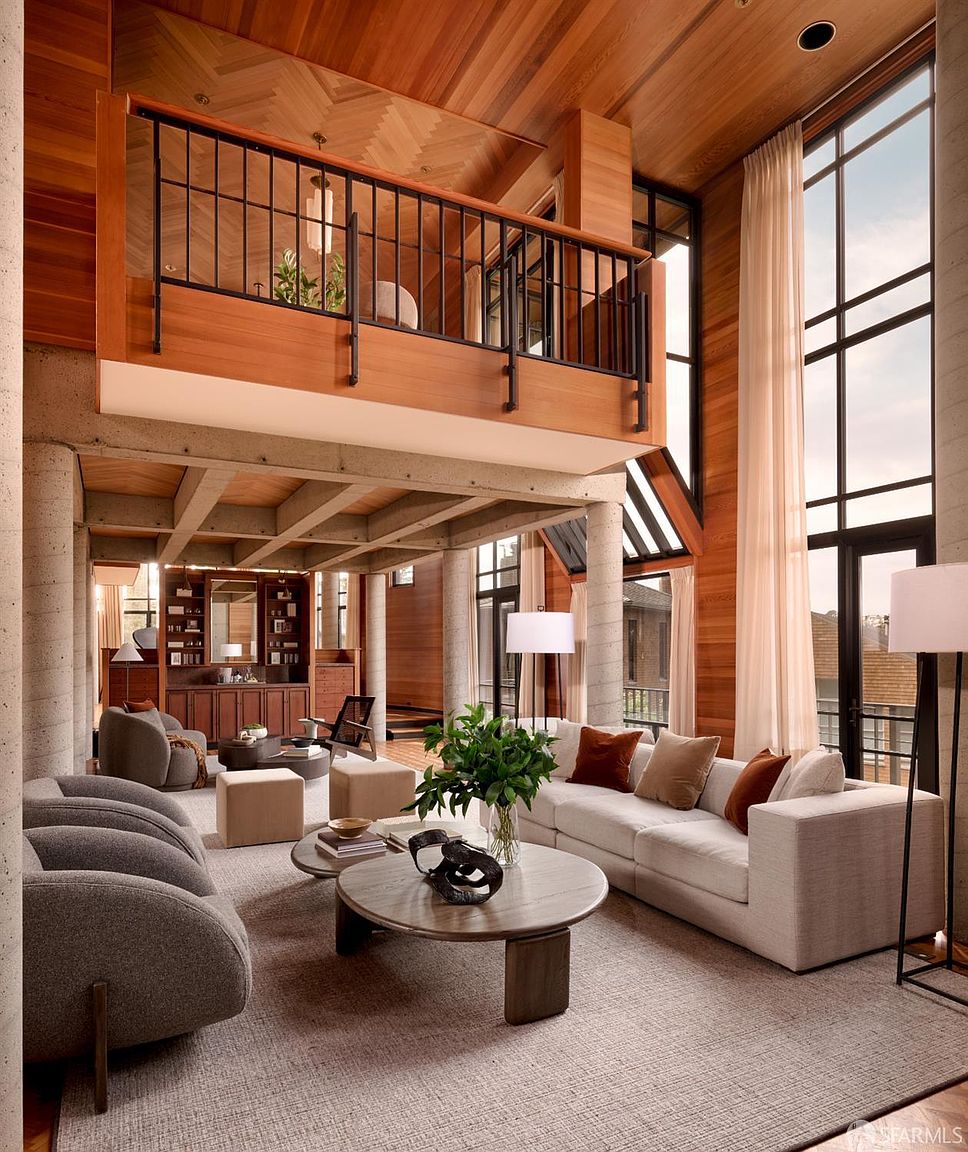 This is a spacious living room featuring a modern design with high ceilings and large windows that provide ample natural light. The room includes a comfortable sofa, armchairs, a round coffee table, and a balcony overlooking the living area. The architectural details, such as the exposed concrete pillars and wooden accents, add to the room's unique character and upscale feel.