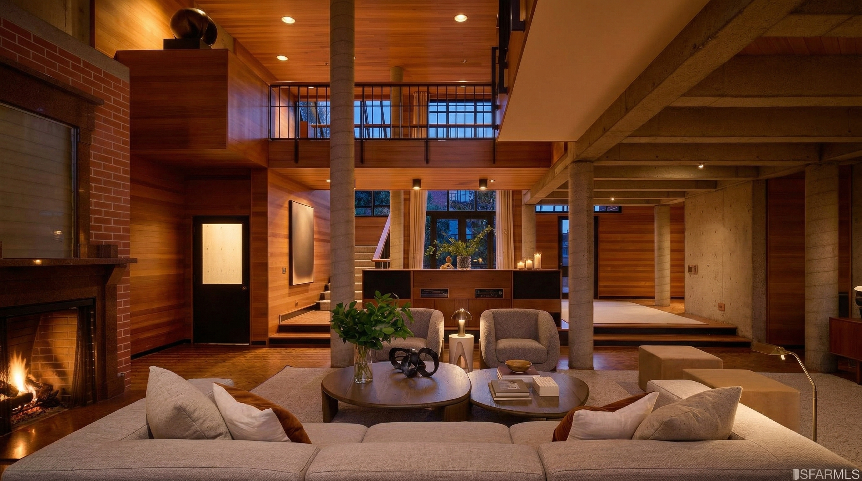 This is an interior shot of a spacious living room featuring a modern design with wood and concrete elements. The room includes a large sectional sofa, round coffee tables, and comfortable armchairs. A unique architectural design is showcased with exposed concrete pillars and wooden walls, creating a warm and inviting atmosphere.