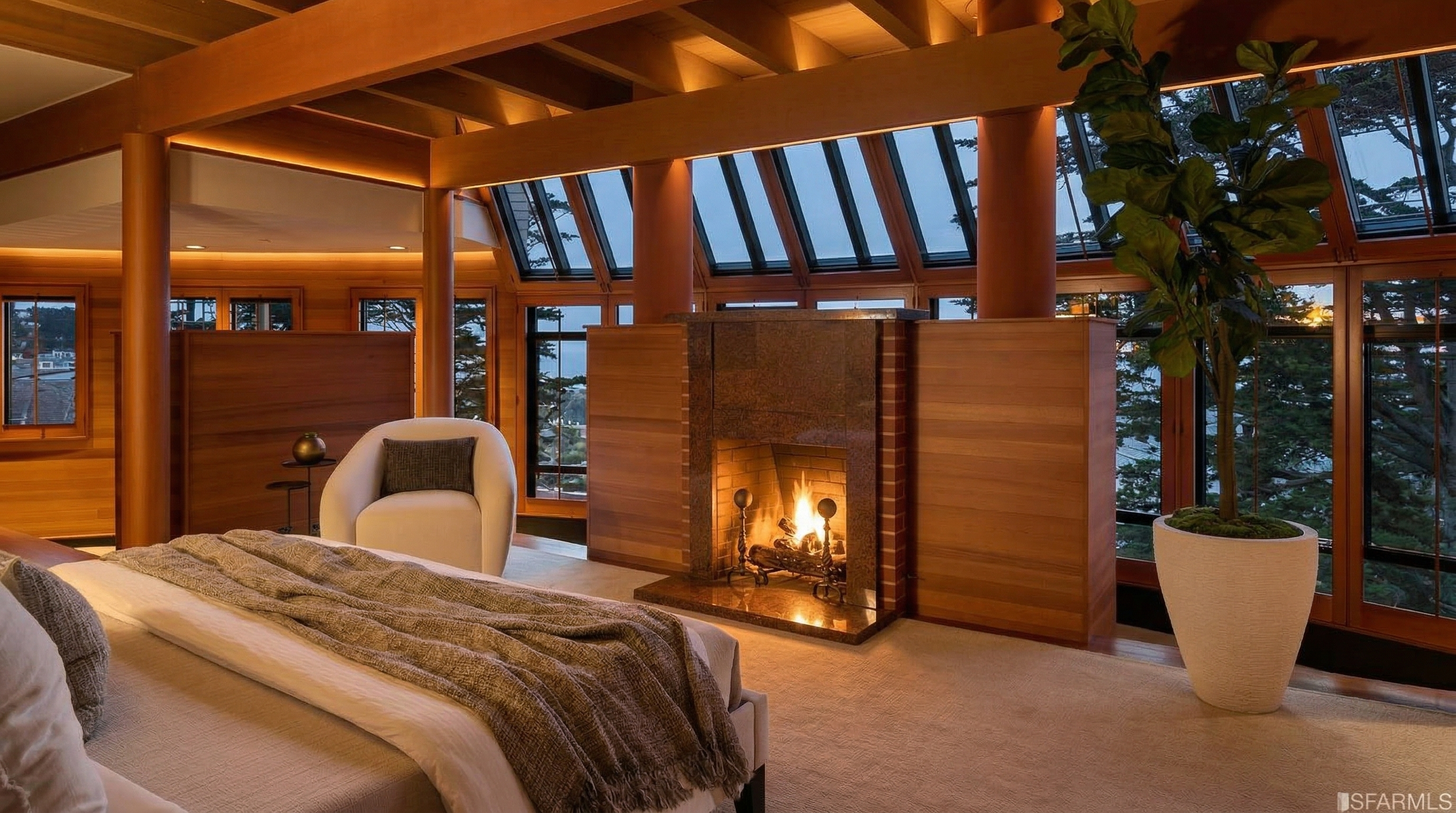 This is a primary bedroom featuring a bed with a throw blanket, a white chair, and a fireplace. The room has wood paneling and beams, with windows that have blinds. A large potted plant sits near the window, adding a touch of nature to the space.