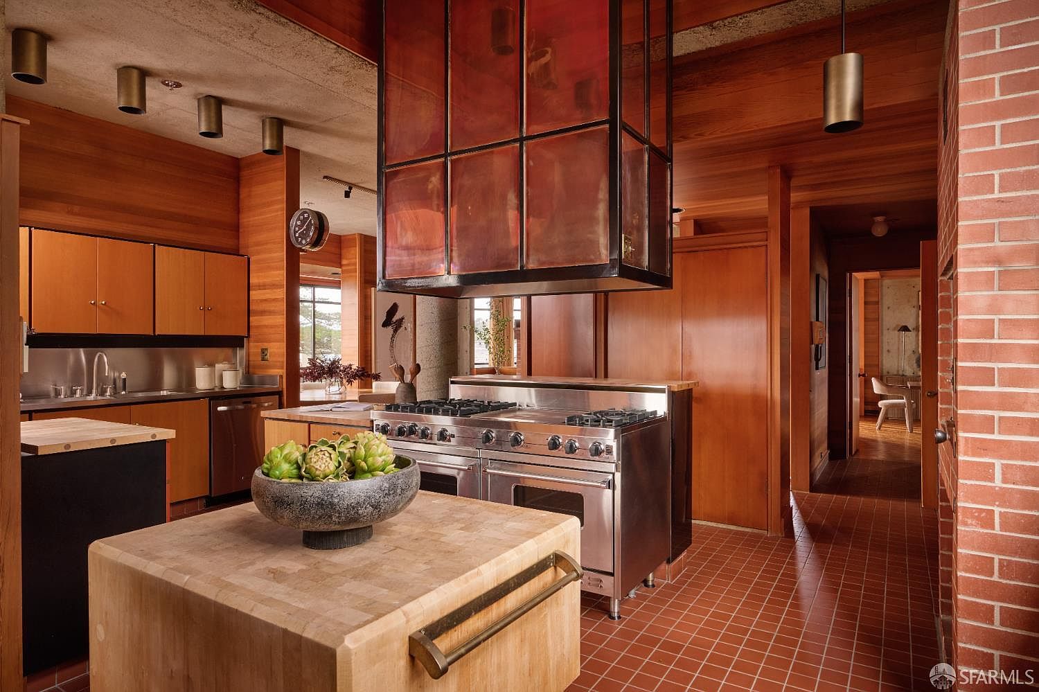 This is a warm and inviting kitchen featuring wood cabinetry, stainless steel appliances, and a unique copper-toned range hood. The kitchen island is topped with a butcher block and a bowl of artichokes, adding a touch of rustic charm. The red brick accent wall and tiled floor contribute to the kitchen's distinctive character, creating a cozy and stylish space.