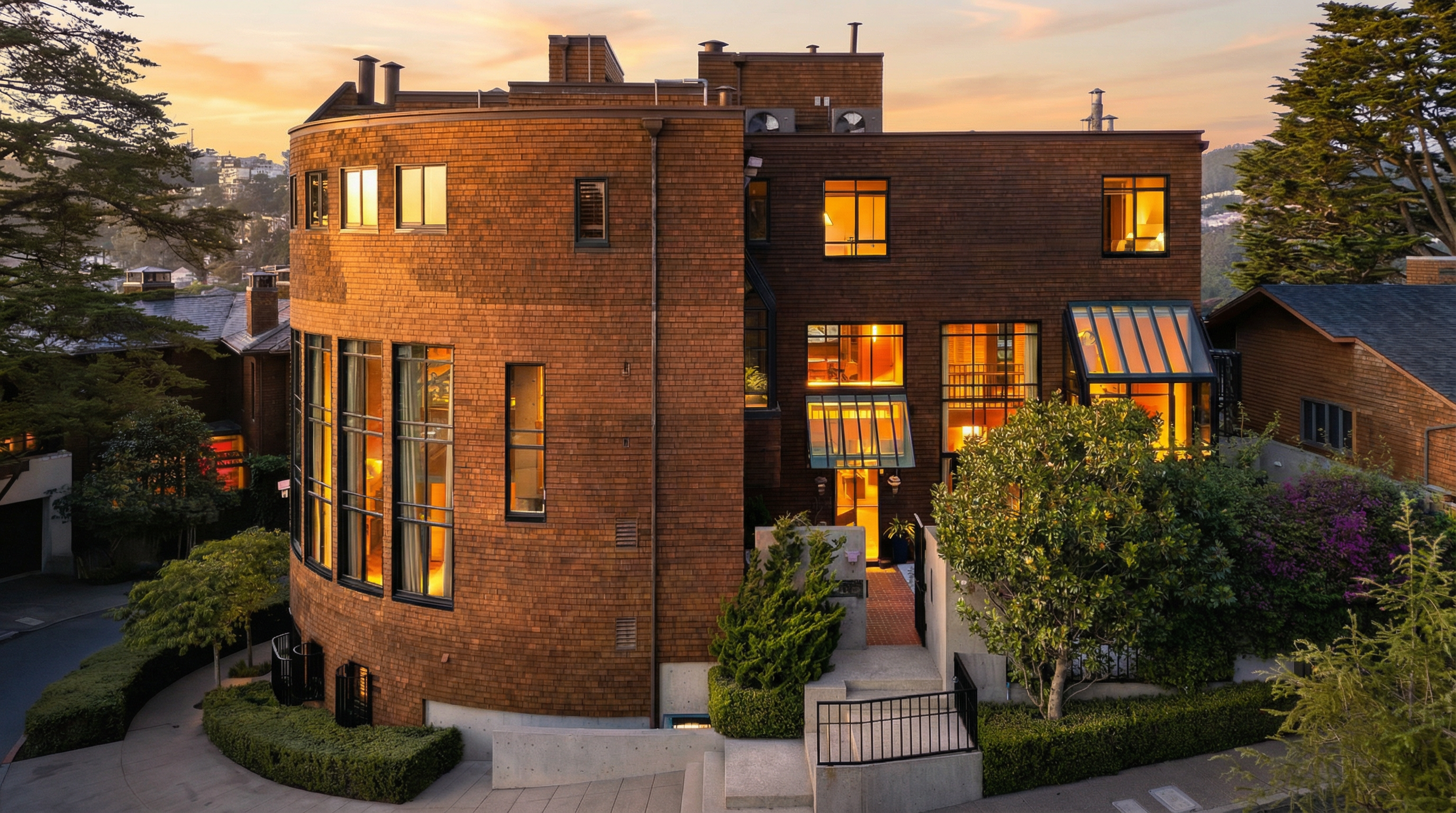 This is a front exterior view of a unique, multi-level home with a rounded section clad in brown shingles. The building features numerous windows that glow with warm interior light, contrasting with the twilight sky. Landscaping includes manicured hedges and trees, adding to the property's curb appeal.