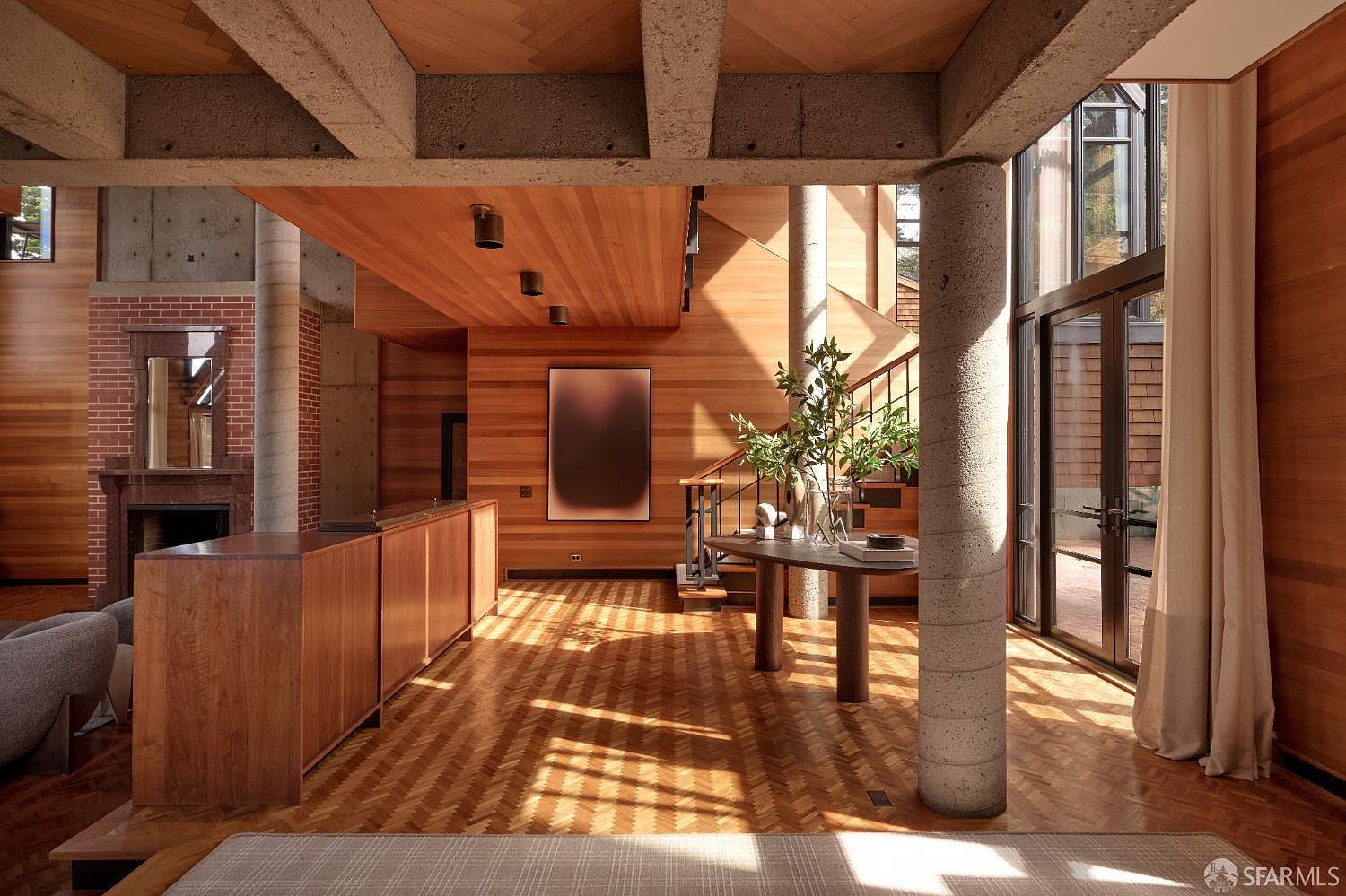 This interior shot showcases a spacious living room with a unique architectural design, featuring exposed concrete beams, warm wood paneling, and herringbone-patterned hardwood floors. A striking concrete pillar anchors the space, while a round table with a plant adds a touch of nature. The room is bathed in natural light, creating a welcoming and sophisticated atmosphere.