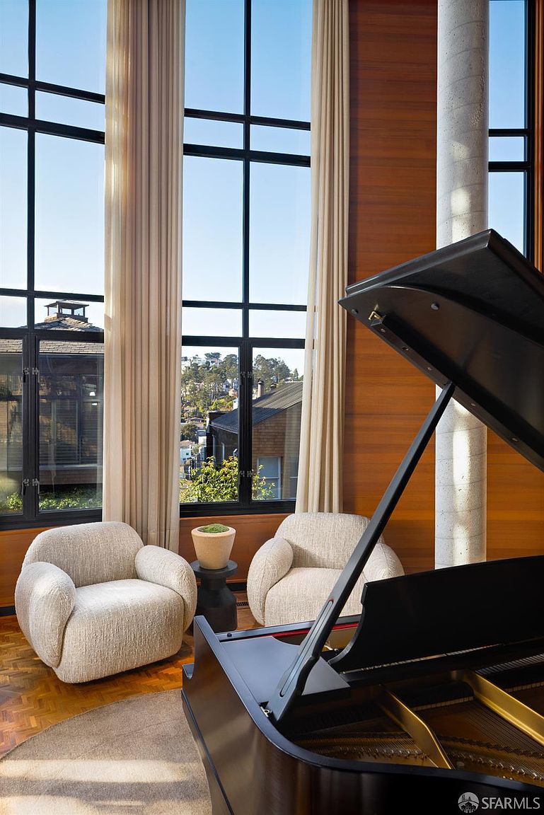 This interior shot showcases a sophisticated living room with floor-to-ceiling windows offering a scenic view. Two plush armchairs flank a small side table, while a grand piano dominates the foreground, adding a touch of elegance and musicality to the space. The room is bathed in natural light, highlighting the warm wood paneling and creating an inviting atmosphere.