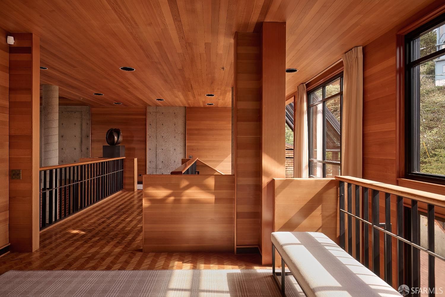 This interior shot showcases a hallway and staircase area with extensive use of wood paneling on the walls and ceiling, creating a warm and inviting atmosphere. The design incorporates concrete elements and black metal railings, adding a touch of modern contrast. Natural light streams in through large windows, highlighting the architectural details and the light-colored bench.