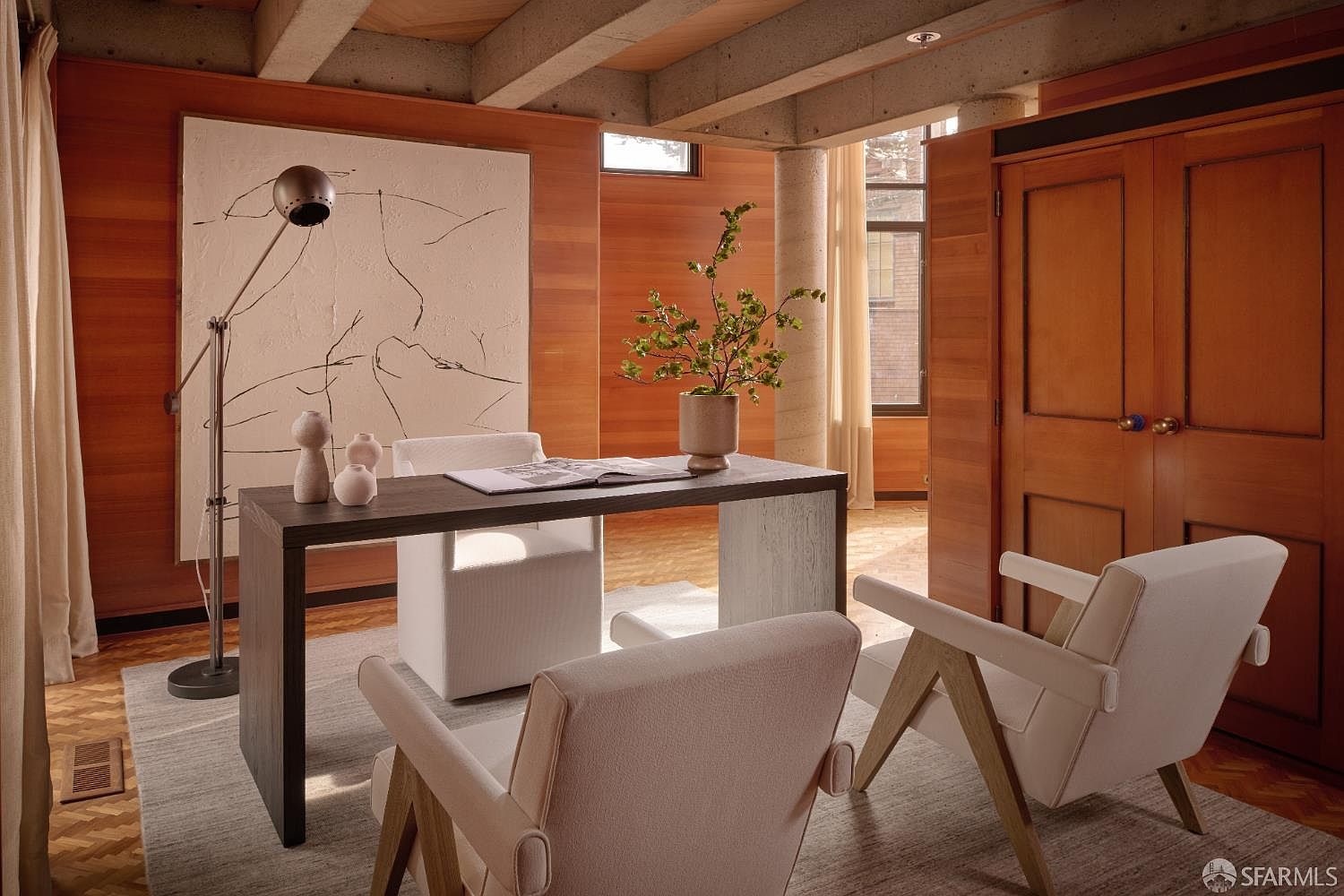 This interior shot showcases a stylish office or study area with warm wood paneling and concrete beam ceilings. A modern desk is adorned with decorative objects and a plant, complemented by two armchairs and a floor lamp. The room exudes a sophisticated and comfortable atmosphere, ideal for a home office setting.
