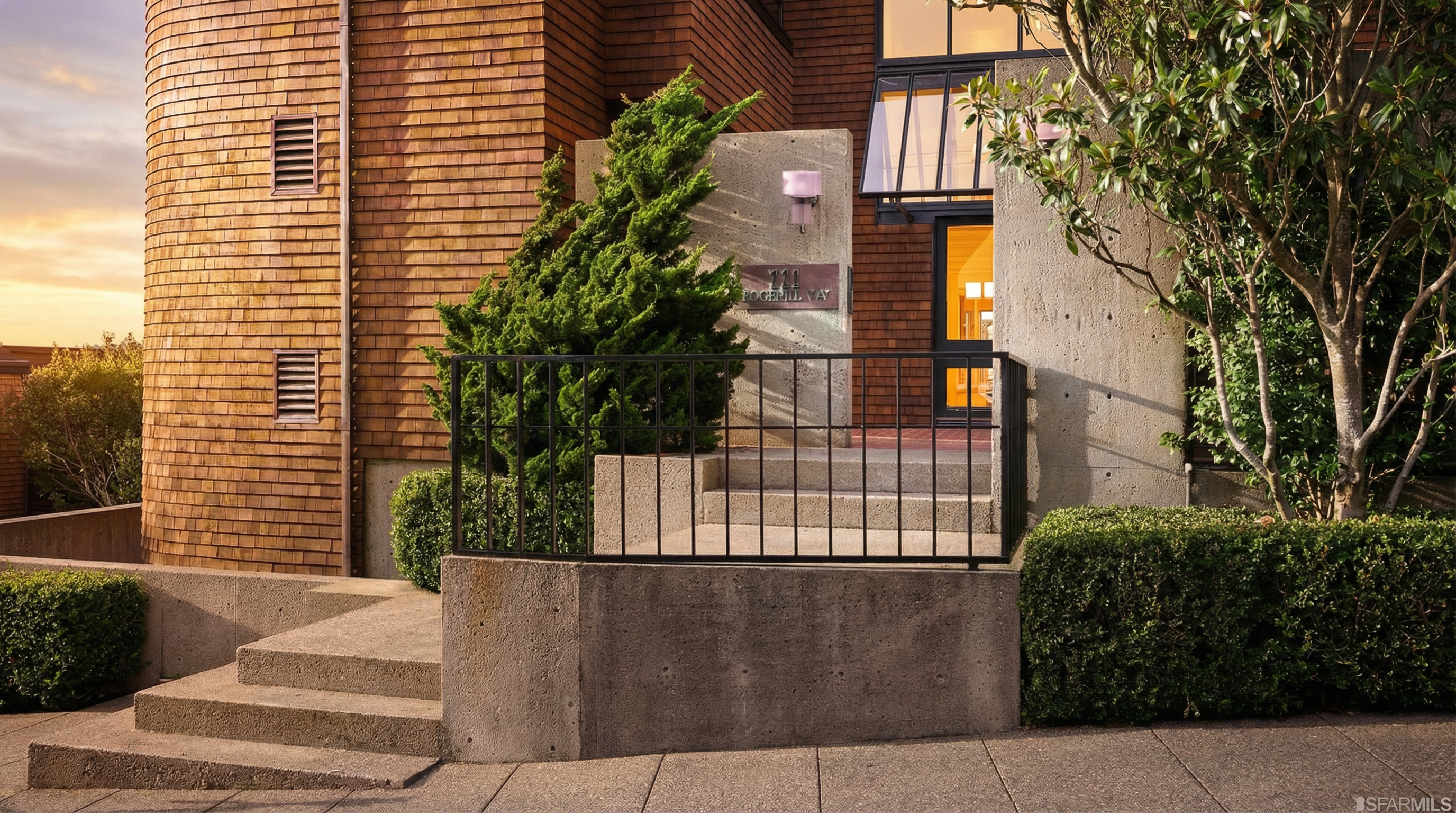 This image showcases the entryway of a building, featuring a concrete staircase leading to the entrance. A black metal railing borders the steps, and greenery flanks both sides of the staircase. The building's facade is a mix of wood shingles and concrete, with a modern architectural style.