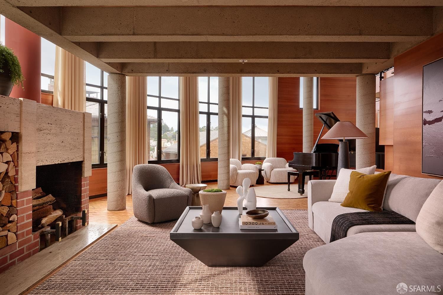 This is a spacious living room featuring a modern design with exposed concrete beams and columns. The room includes a fireplace, a grand piano, and comfortable seating arrangements, all illuminated by natural light streaming through large windows. The overall impression is one of sophisticated comfort and architectural interest.
