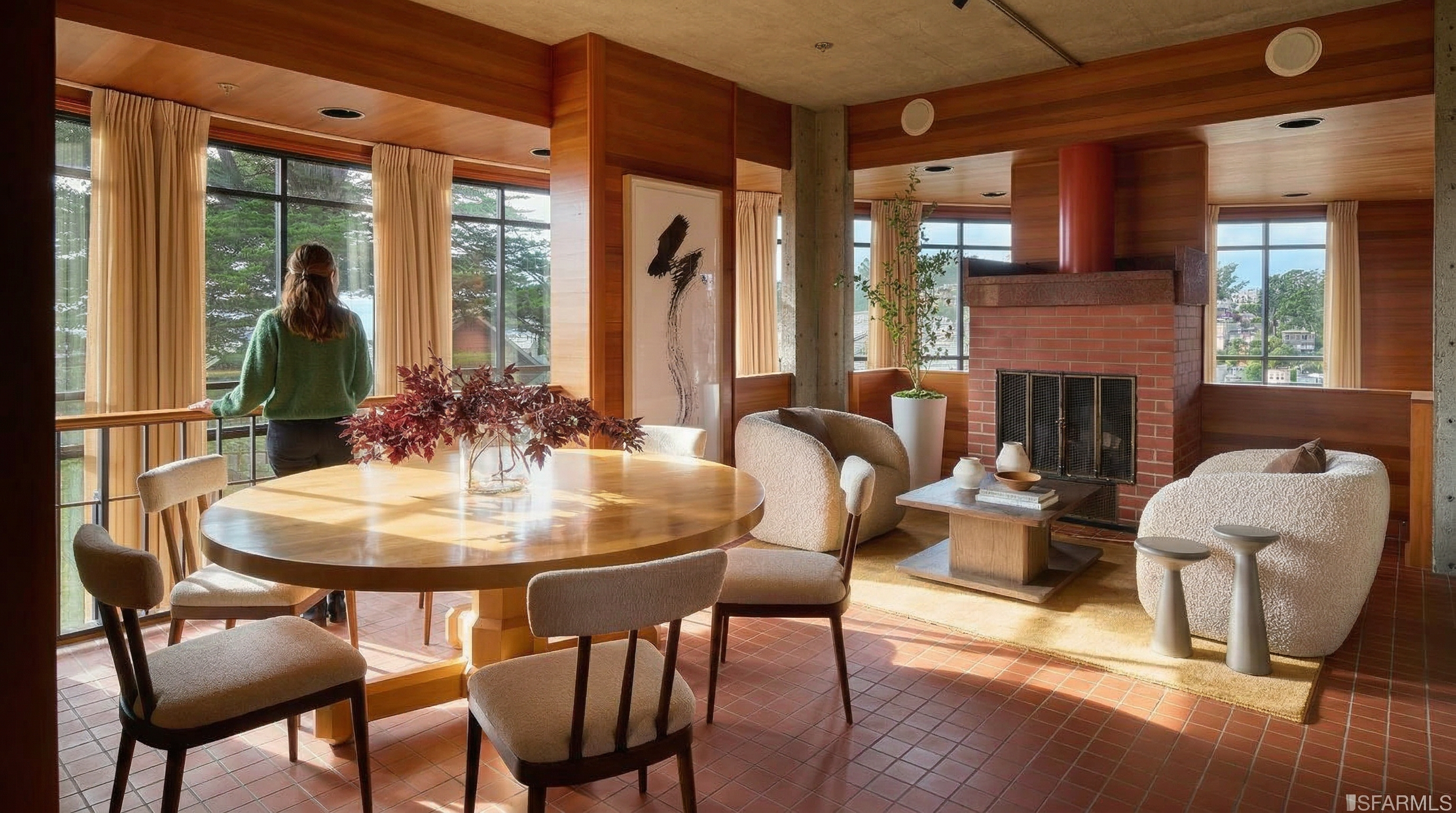 This interior shot showcases a dining room with a warm and inviting atmosphere. A round wooden table is surrounded by chairs with neutral upholstery, complemented by a vase of dark red foliage as a centerpiece. The room features a brick fireplace, comfortable armchairs, and large windows that offer natural light and views of the outdoors, creating a cozy and sophisticated space.