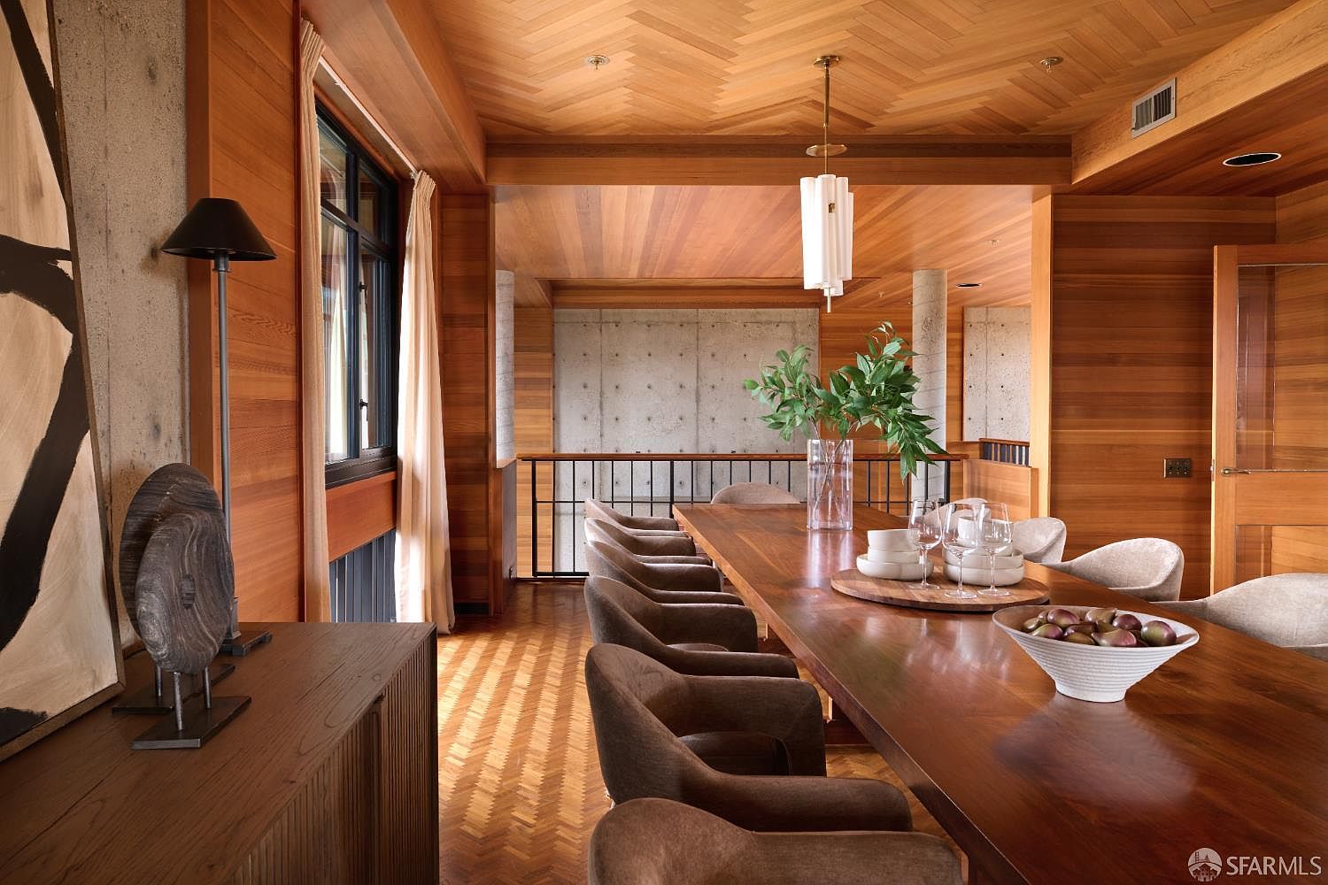 This is an interior shot of a dining room featuring a long wooden table with brown upholstered chairs. The room has wood-paneled walls and a herringbone wood floor pattern. A modern light fixture hangs above the table, and there are decorative items such as a vase with greenery and bowls on the table. The room has a warm and inviting atmosphere.