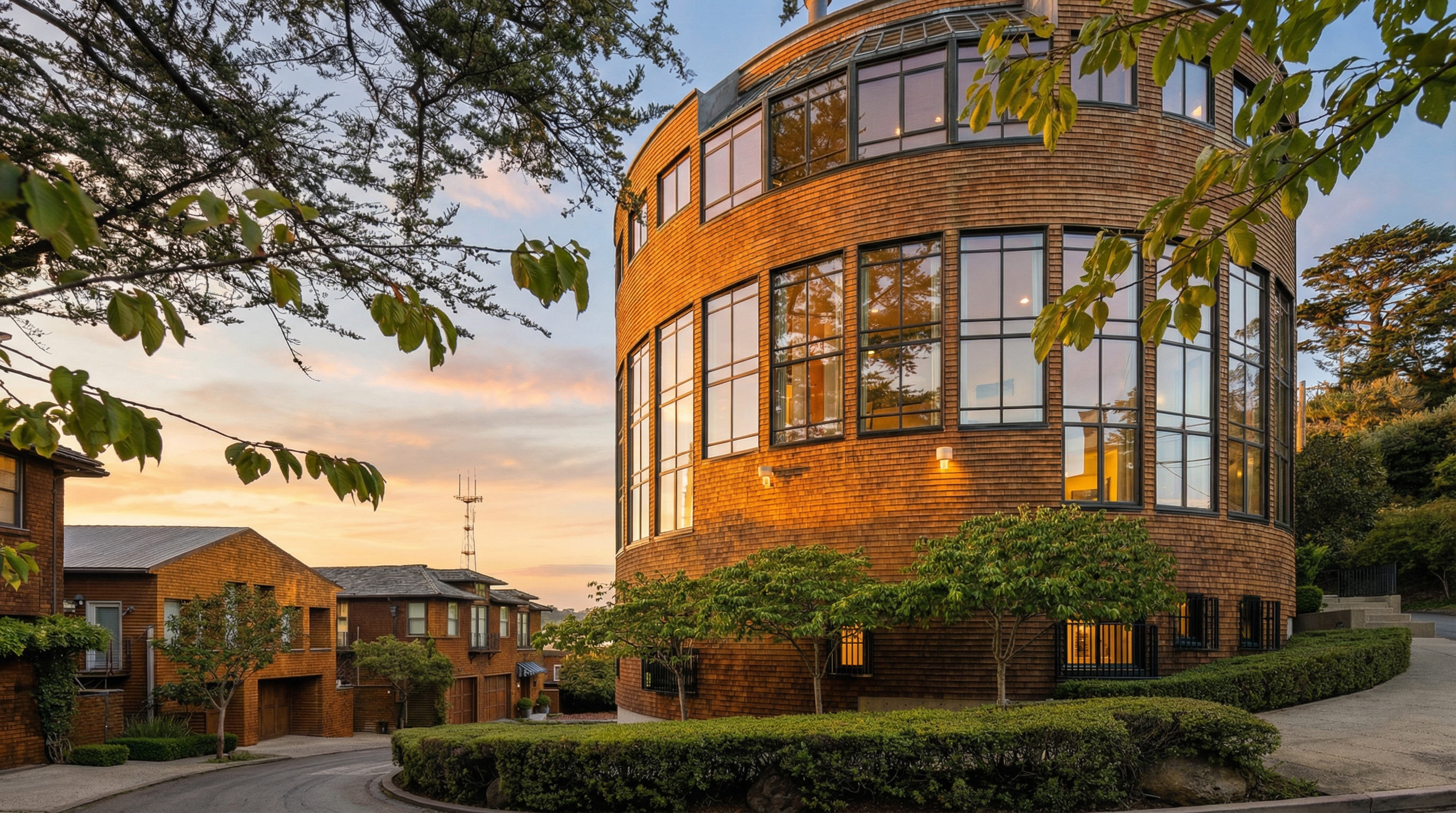This image showcases the front exterior of a unique, cylindrical building with a brick facade and large windows. The building is surrounded by manicured greenery and a circular driveway. Adjacent to the main structure are traditional brick townhouses, creating a blend of architectural styles.