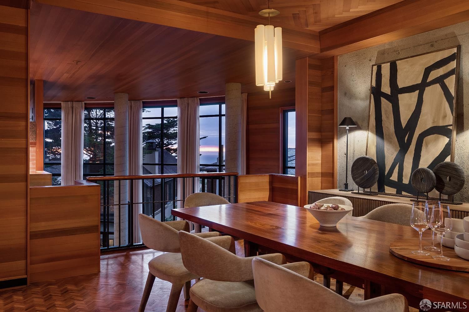 This is an interior shot of a dining room featuring a large wooden table surrounded by upholstered chairs. The room has wood-paneled walls and ceiling, with large windows offering a view of the outside. A modern light fixture hangs above the table, and artwork is displayed on the wall, creating a sophisticated and inviting atmosphere.