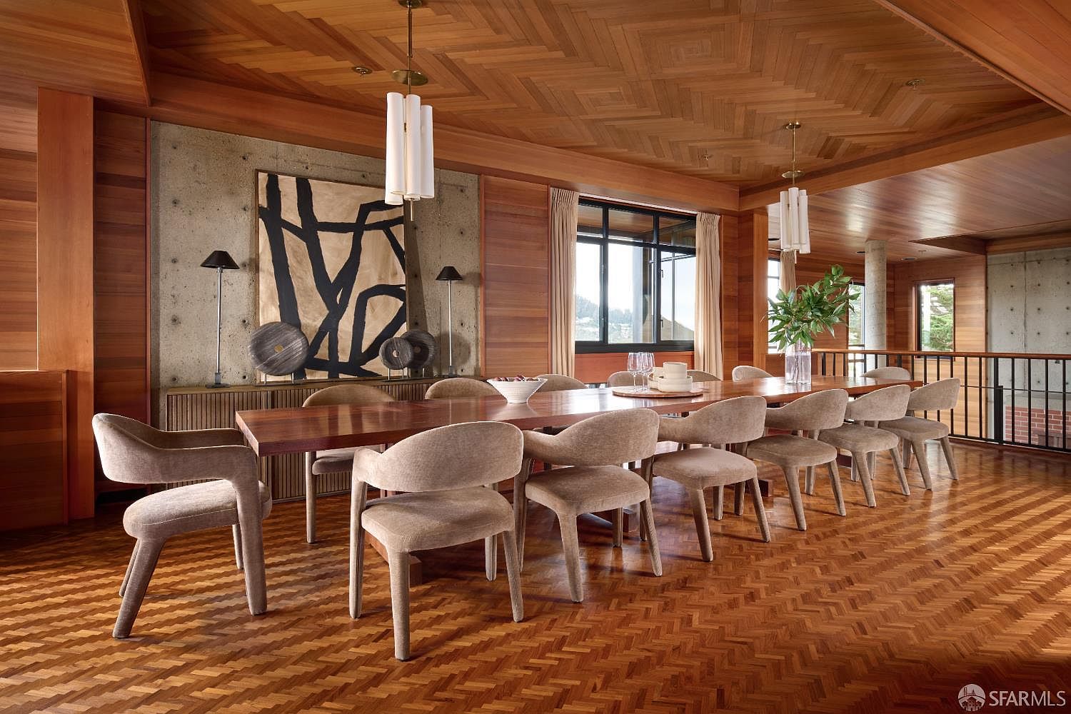 This is an interior shot of a dining room featuring a long wooden table surrounded by upholstered chairs. The room has a warm, inviting atmosphere with wood paneling on the walls and a unique herringbone pattern on the floor and ceiling. A large abstract painting hangs on the wall, complemented by modern lighting fixtures above the table.