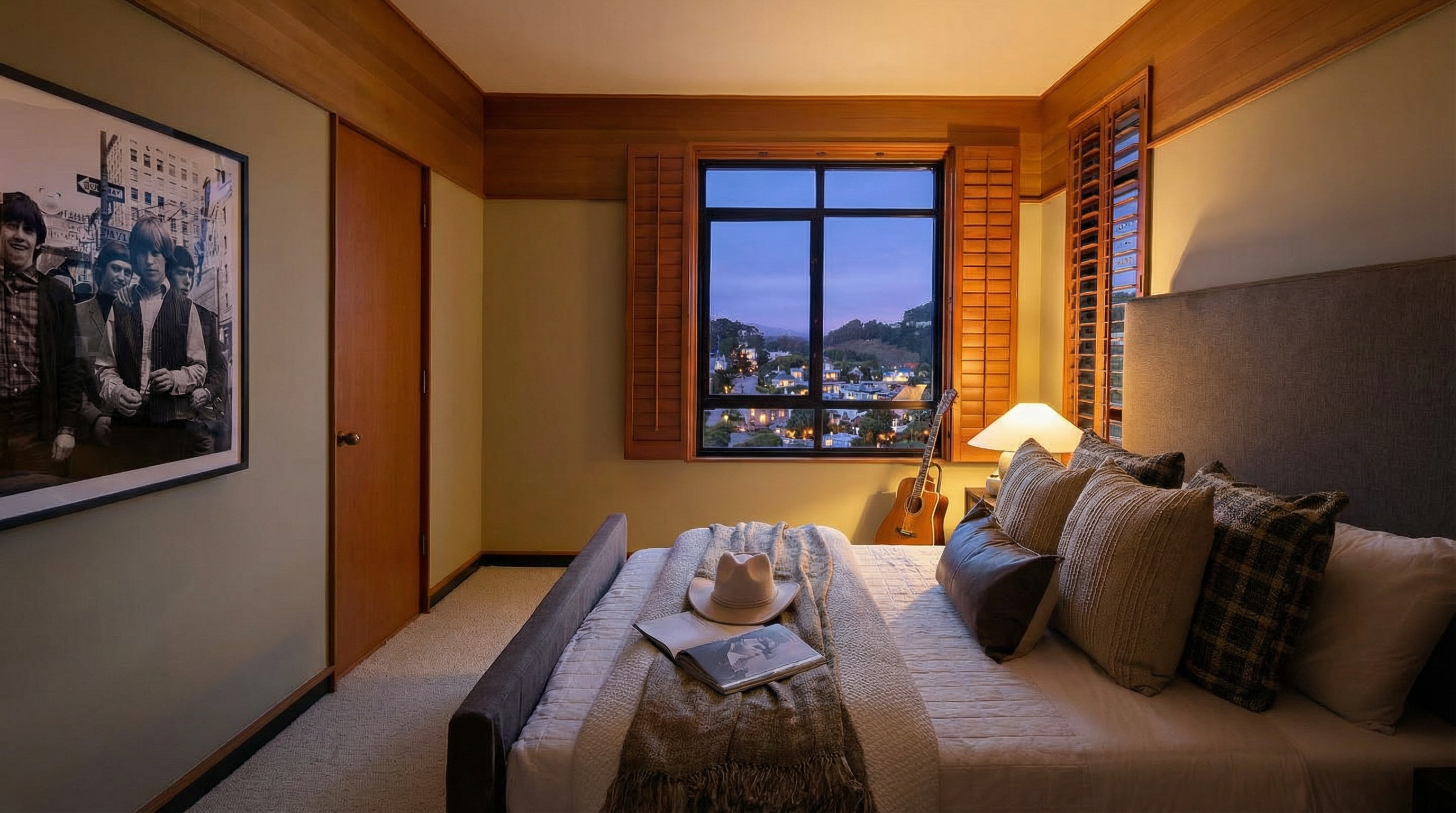 This is a cozy bedroom featuring a bed with multiple pillows and a throw blanket, a window with a view of a cityscape, and a framed black and white photograph on the wall. The room has a warm and inviting atmosphere, with wood trim and neutral wall colors. The bed is neatly made with a white comforter and several decorative pillows.