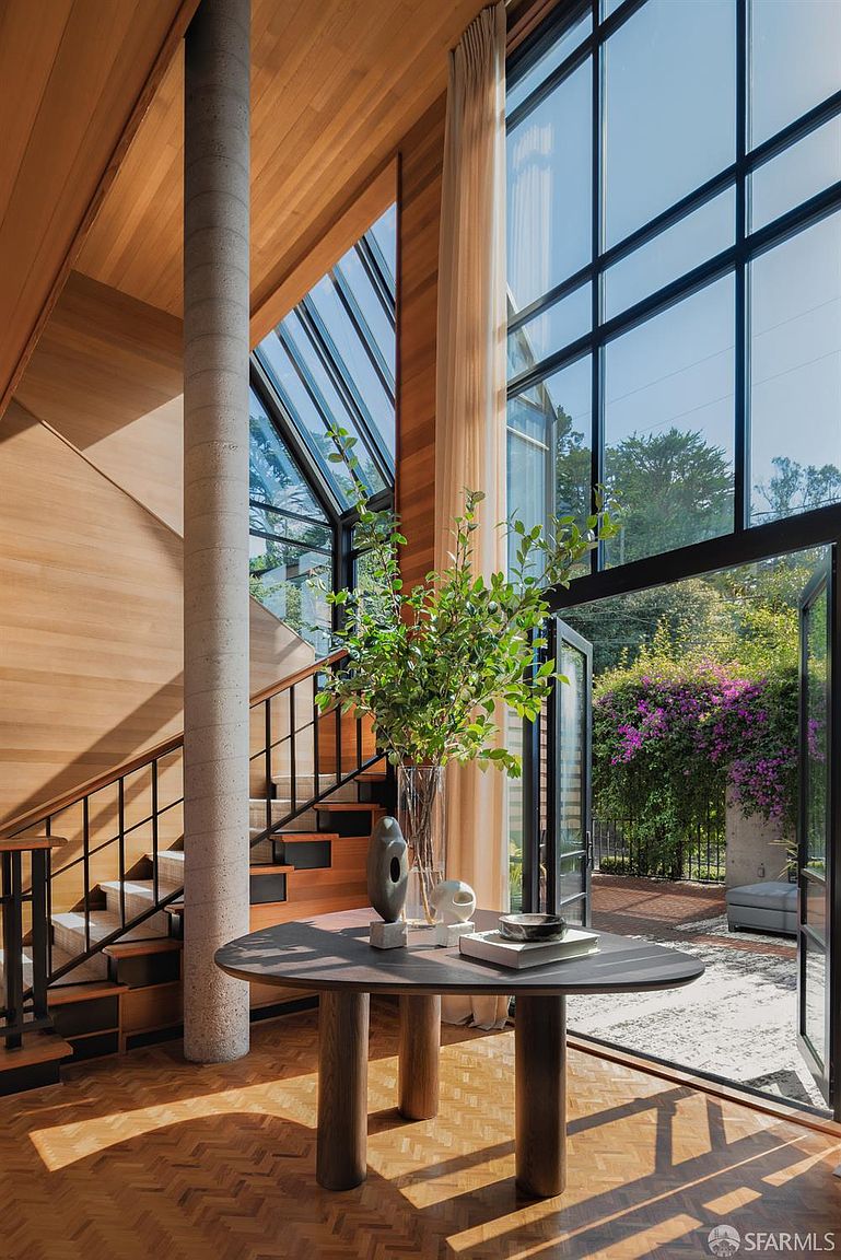This interior shot showcases a modern hallway with a staircase, featuring wood paneling, a concrete pillar, and large windows that bring in natural light. A round table with decorative items and a vase of greenery adds a touch of elegance, while the open doorway leads to an outdoor garden, creating a seamless transition between indoor and outdoor spaces. The perspective is from a low angle, emphasizing the height and architectural details of the space.