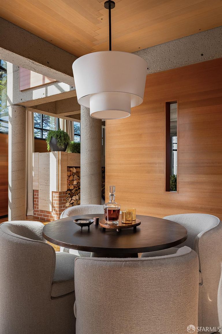 This interior shot showcases a dining area with a round dark wood table surrounded by four upholstered chairs. A modern, tiered white pendant light hangs above the table, and a wooden wall with a vertical window adds a touch of warmth. The room features concrete columns and a glimpse of an adjacent space with a fireplace, creating a sophisticated and inviting atmosphere.