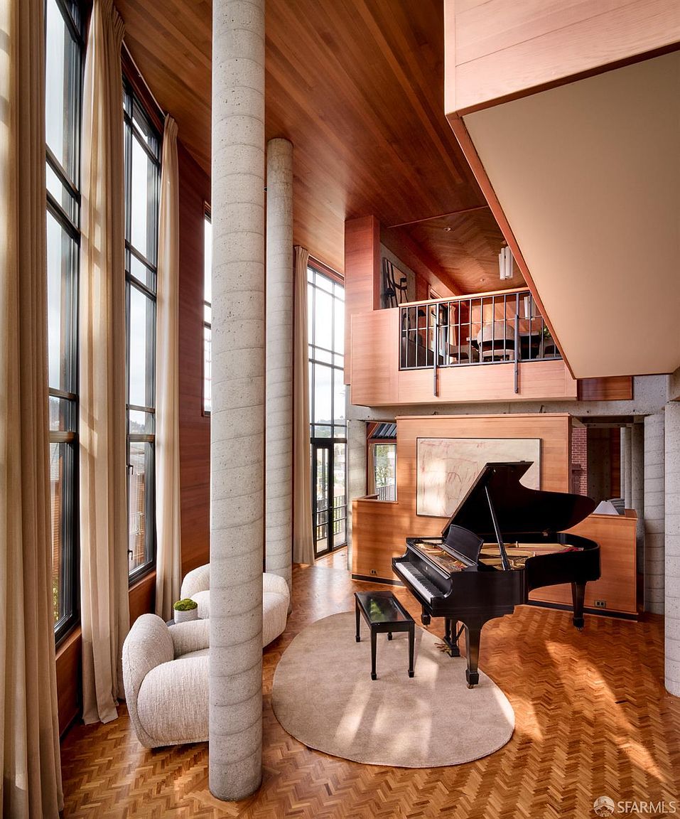 This grand living room features soaring ceilings, floor-to-ceiling windows draped with elegant curtains, and striking architectural columns. A black grand piano sits on a circular rug, adding a touch of sophistication to the space. The warm wood tones of the ceiling and floors create an inviting atmosphere, while the balcony overlooking the room adds a unique architectural element.