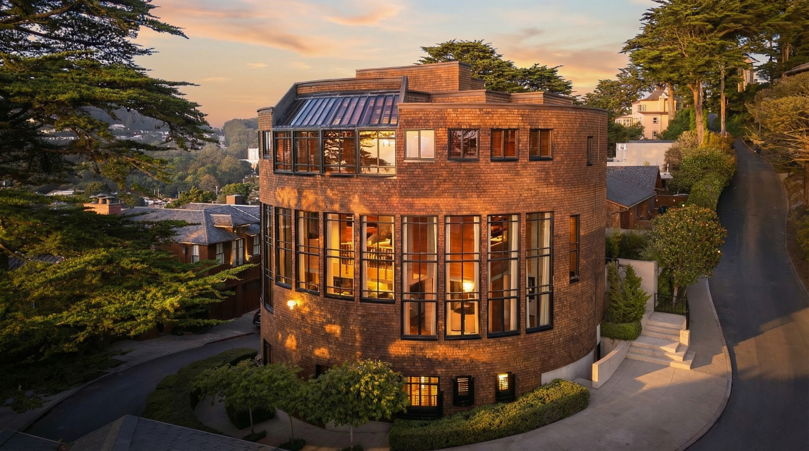 This striking, cylindrical architectural landmark is clad in warm-toned shingles and features extensive floor-to-ceiling glass windows that wrap around the curvature of the structure. Captured at golden hour, the building exudes a sophisticated, modern aesthetic that blends uniquely with the surrounding landscape. Its elevated design and prominent position within the hillside neighborhood offer a compelling mix of architectural distinction and scenic views.