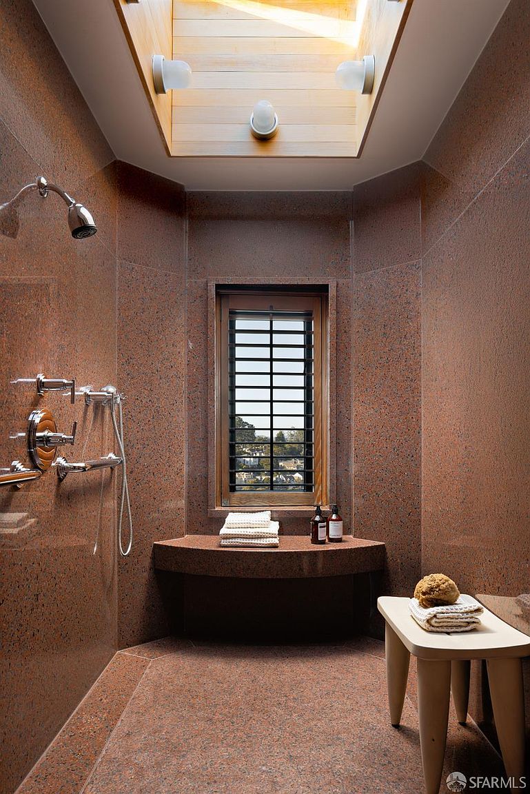 This is a primary bathroom featuring red granite walls and flooring, a built-in bench under a window with blinds, and a shower area with chrome fixtures. A skylight provides natural light, and a small stool with towels and a sponge adds a spa-like touch. The overall impression is luxurious and unique.