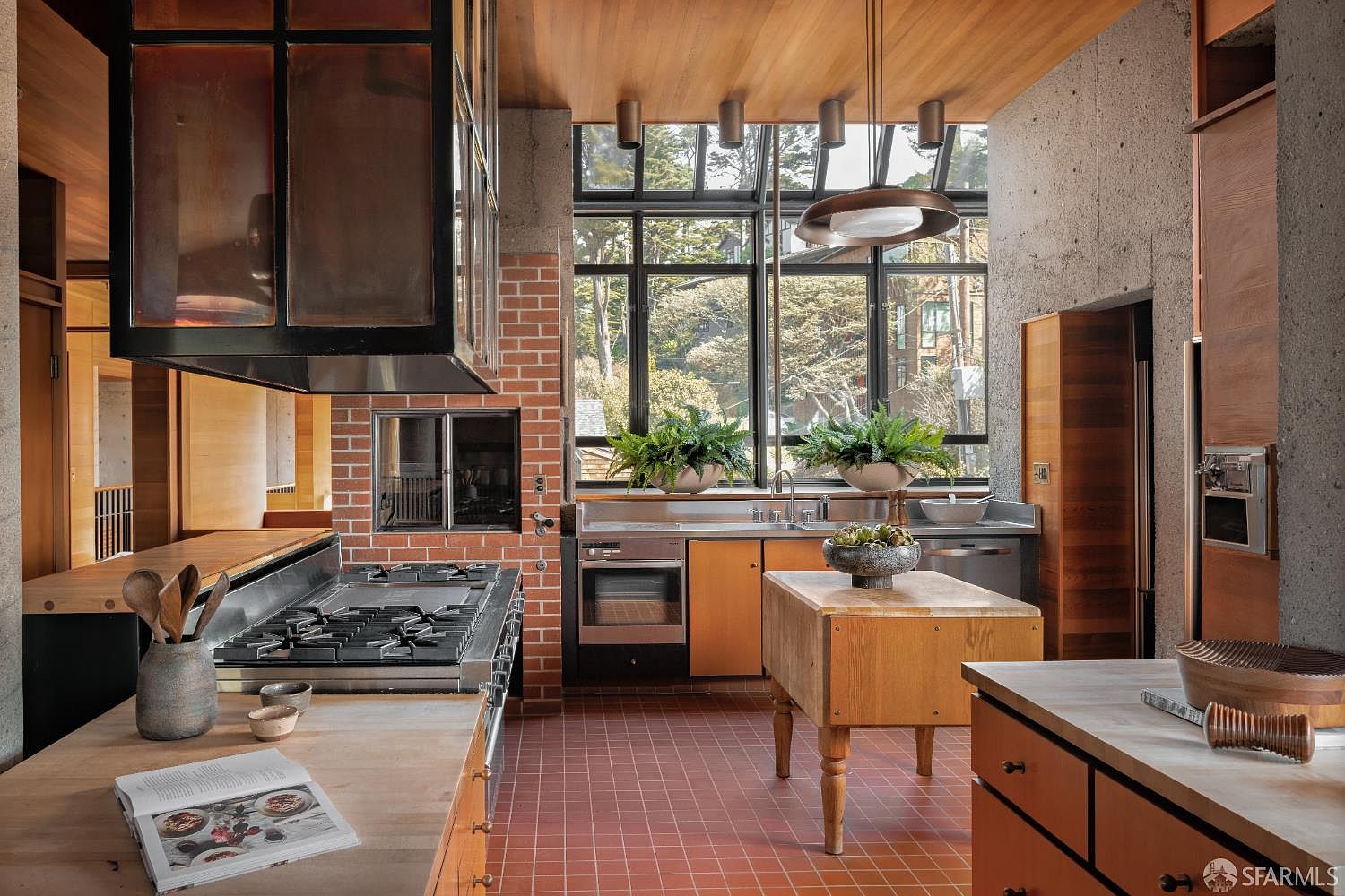 This kitchen features a unique blend of materials, including brick, concrete, and wood, creating a warm and inviting atmosphere. A large window provides ample natural light and views of the outdoors, while the central island offers additional workspace. The stainless steel appliances and custom cabinetry add to the kitchen's functionality and style.
