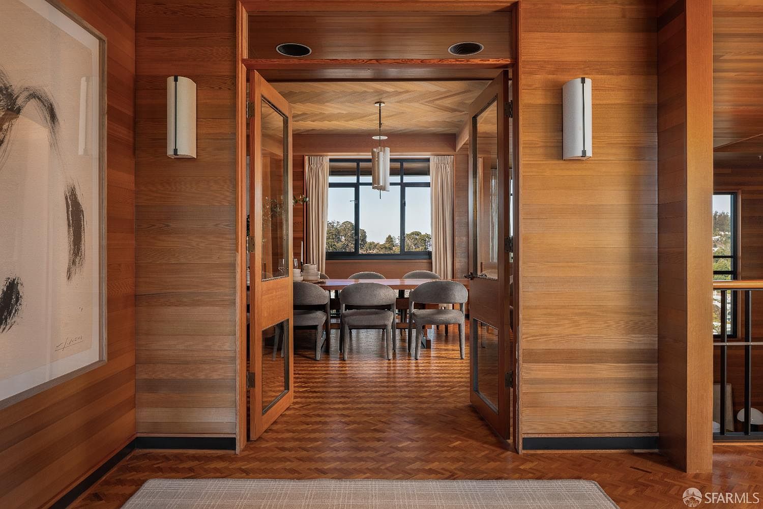 This interior shot showcases a dining room with rich wood paneling and herringbone-patterned floors, creating a warm and sophisticated ambiance. The room is viewed through an open doorway, revealing a dining table with modern chairs and a window offering a glimpse of the outdoors. Symmetrical wall sconces flank the doorway, adding to the room's balanced aesthetic.