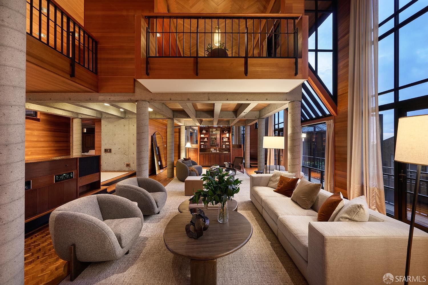 This is a spacious living room featuring a modern design with wood paneling and concrete pillars. The room is furnished with a large sectional sofa, two armchairs, and a round coffee table, all set on a neutral-toned rug. Large windows provide ample natural light, and a balcony overlooks the living area, creating a sense of openness and grandeur.