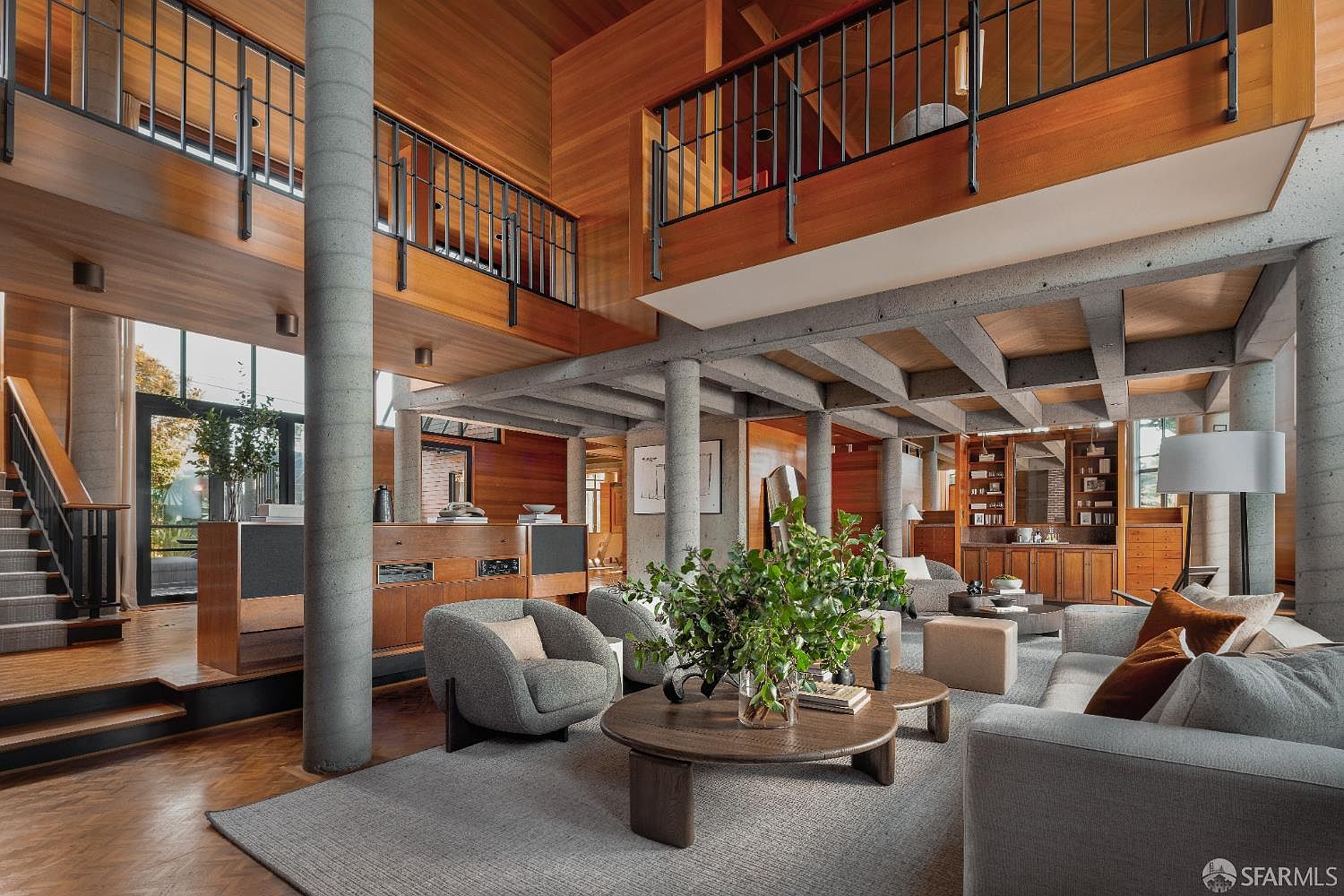 This is a spacious living room with a modern architectural design, featuring exposed concrete beams and columns, warm wood paneling, and a multi-level layout. The room is furnished with comfortable gray sofas and armchairs, centered around a round coffee table with a lush green plant. Large windows provide natural light, and the overall impression is one of sophisticated comfort and contemporary style.