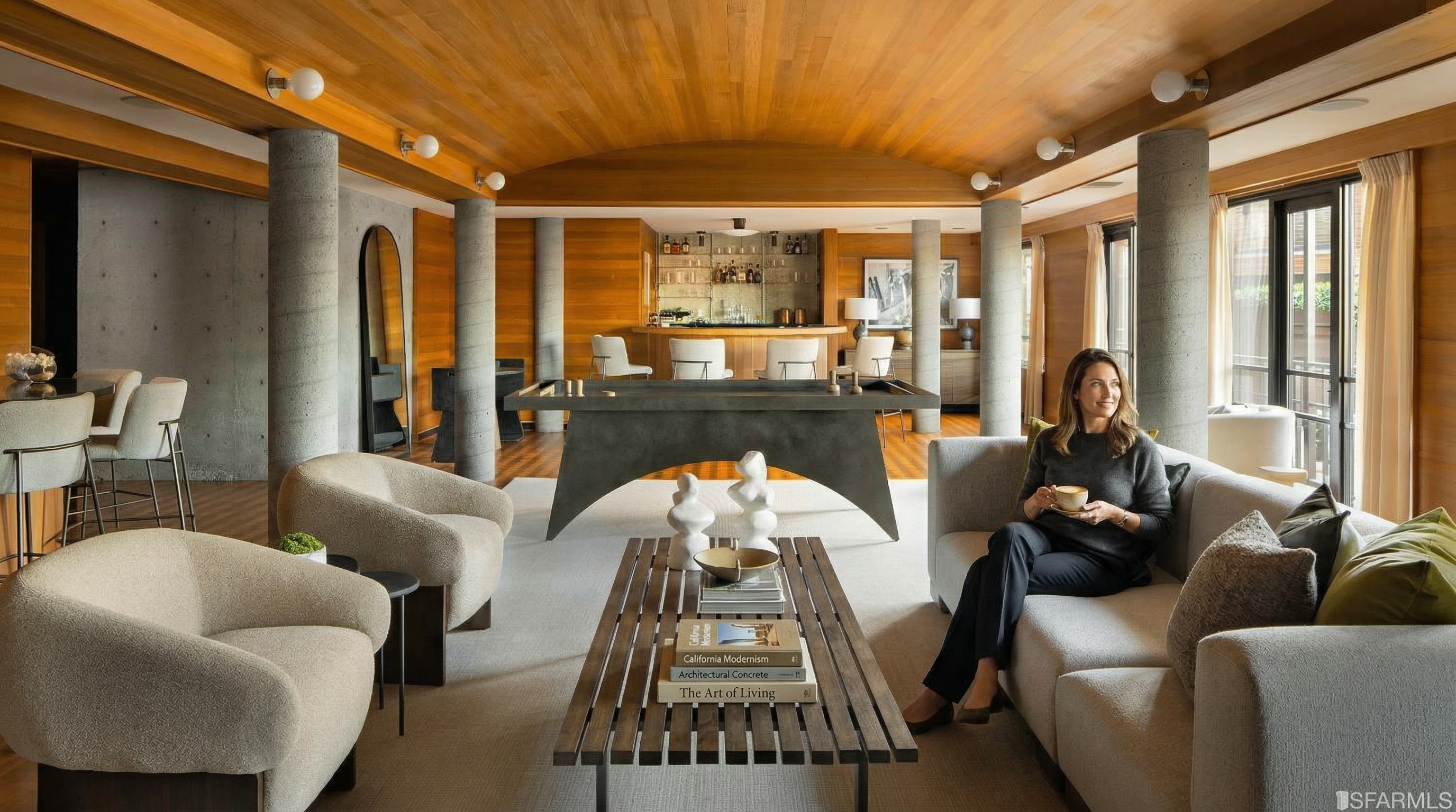 This is an interior shot of a luxurious living room featuring a unique architectural design with concrete pillars and a wooden ceiling. The room is furnished with comfortable seating, including a sofa and armchairs, arranged around a wooden coffee table. A bar area is visible in the background, adding to the room's entertainment appeal.