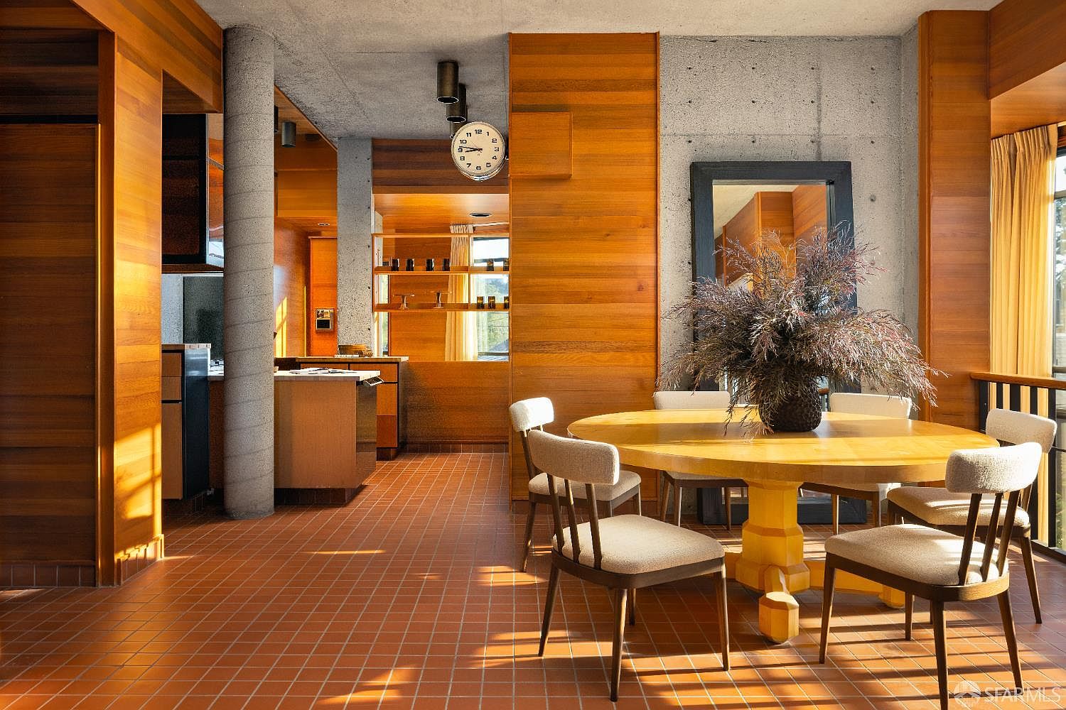 This interior shot showcases a dining room with a mid-century modern aesthetic. The room features a round wooden table surrounded by chairs with light-colored upholstery, set against a backdrop of wood paneling and concrete walls. The warm lighting and natural materials create an inviting and stylish dining space.