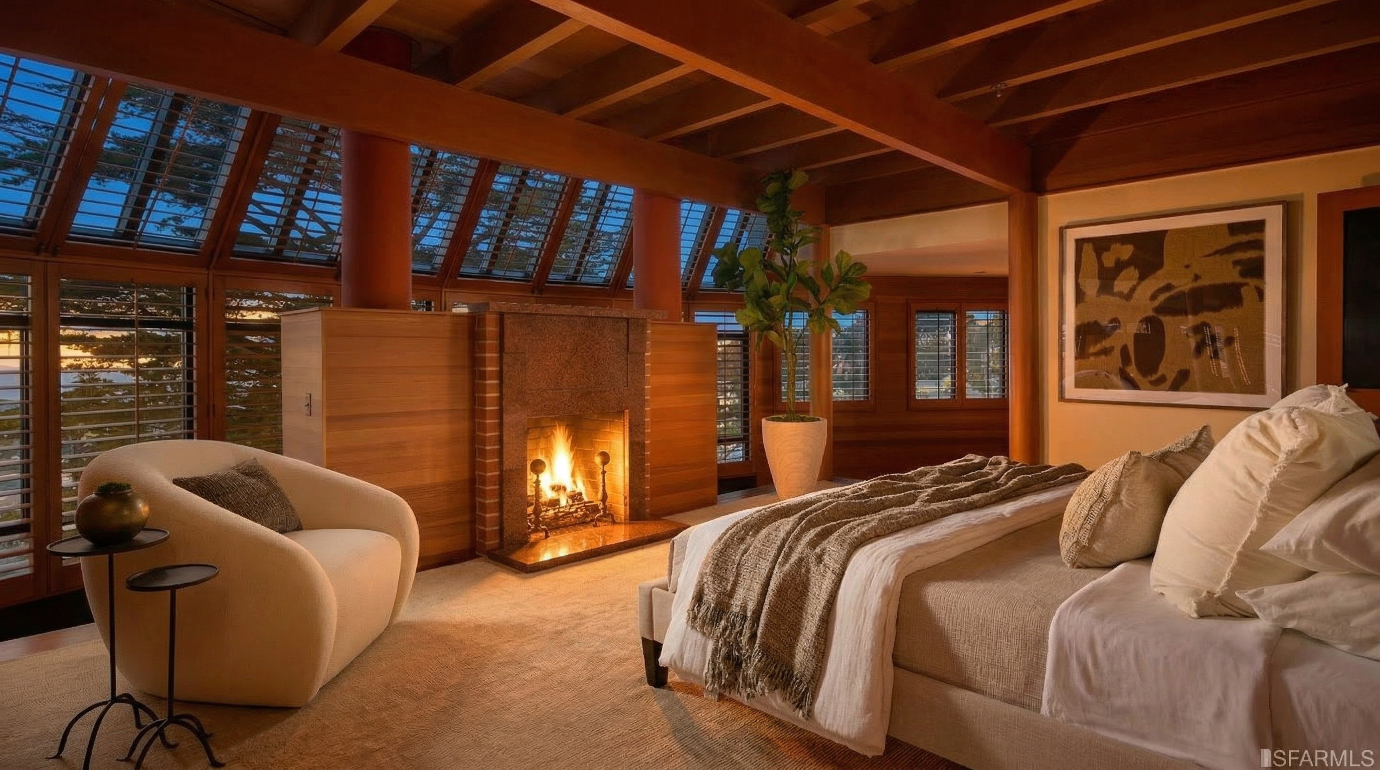 This is a primary bedroom featuring a bed with neutral linens and a throw blanket, a fireplace with a red brick surround, and a comfortable armchair. The room has wood-paneled walls and ceiling beams, and large windows with shutters provide natural light. The overall style is warm and inviting, with a focus on natural materials.
