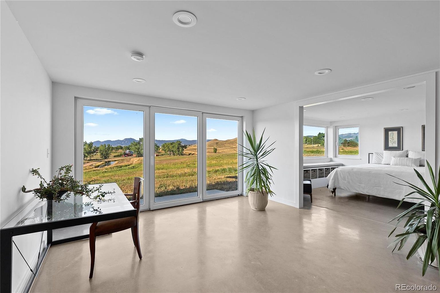 This is a bright and airy primary bedroom with an open floor plan that seamlessly connects to a sitting area. The room features large windows offering scenic views, polished concrete floors, and minimalist decor. The style is modern and serene, emphasizing natural light and a connection to the outdoors.
