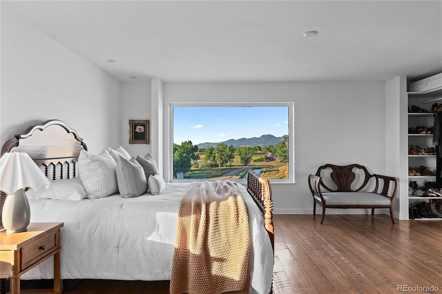 This is a bright and airy primary bedroom featuring a large window with a scenic view of a rural landscape. The room is decorated in a neutral color palette with white walls and bedding, complemented by wooden furniture and flooring. A cozy throw blanket adds a touch of warmth to the space.