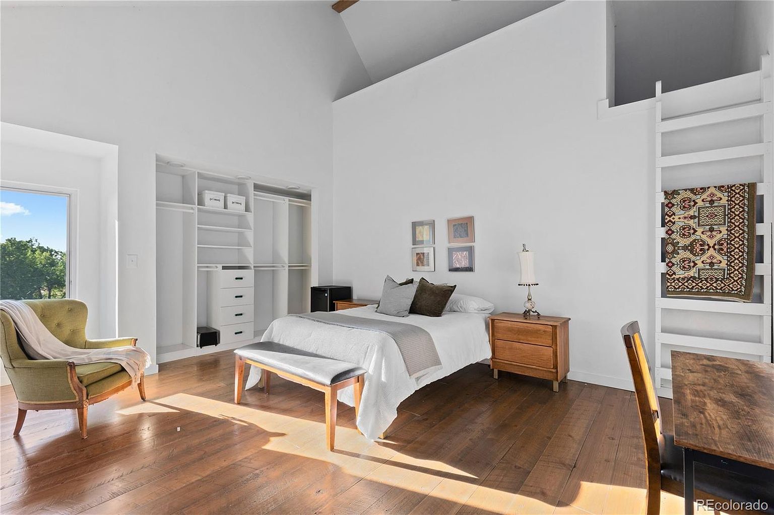 This is a bright and airy primary bedroom featuring hardwood floors, white walls, and a high vaulted ceiling. The room includes a bed with neutral bedding, a bench, a nightstand with a lamp, and a built-in closet system. Natural light floods the space, creating a warm and inviting atmosphere.