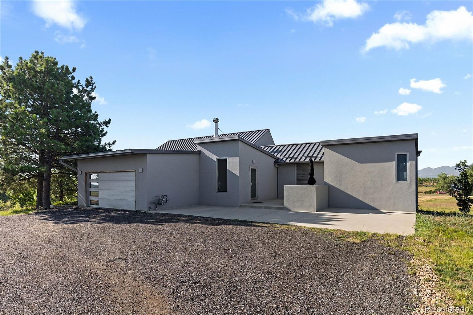 This is a front exterior view of a modern, single-story home with a gray stucco finish and a dark metal roof. The house features a minimalist design with clean lines, a garage on the left, and a concrete driveway leading up to the entrance. The landscape includes a gravel area in the foreground and some natural vegetation in the background, under a partly cloudy sky.