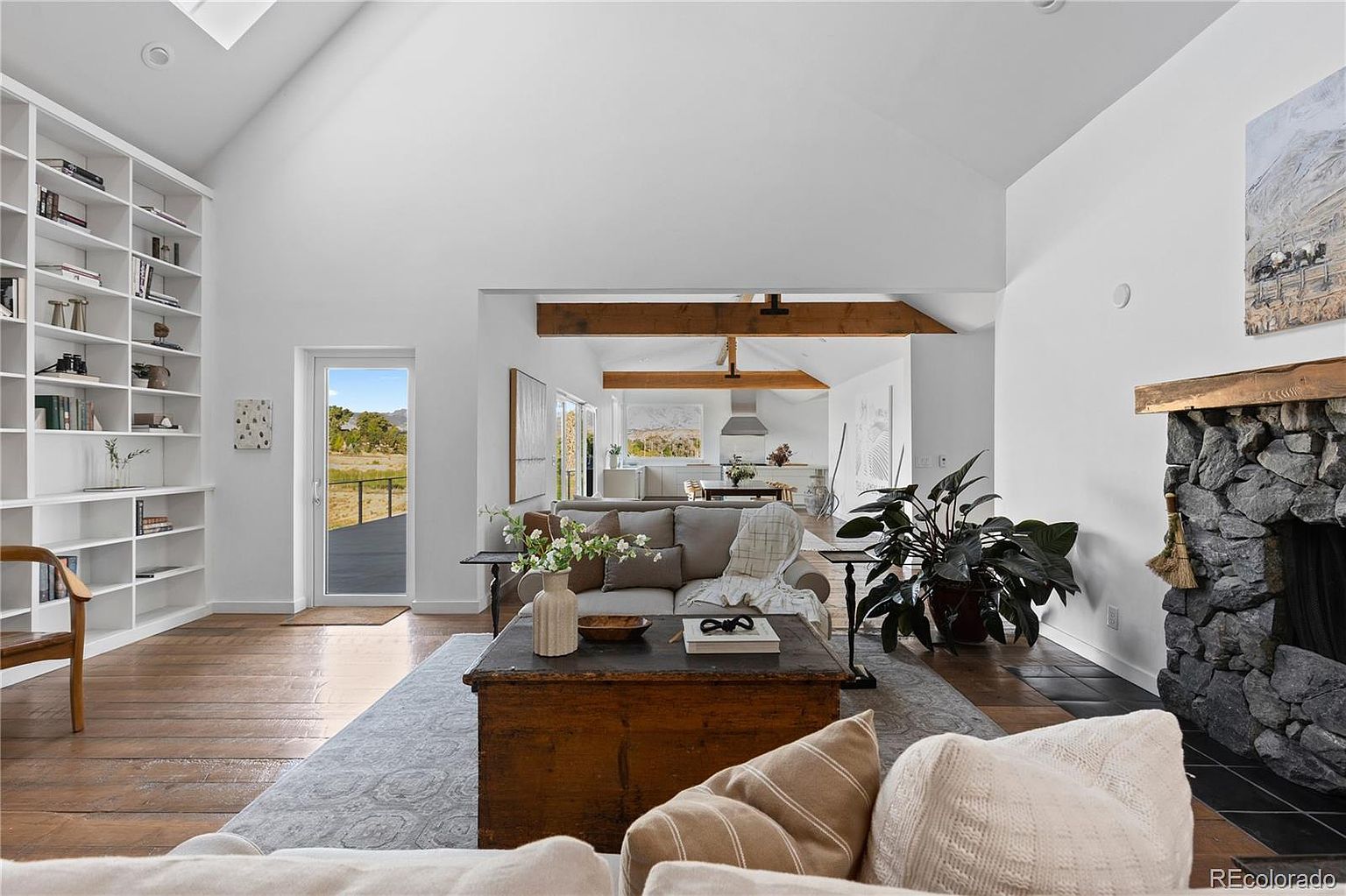 This is an interior shot of a living room featuring a stone fireplace, a wooden coffee table, and a comfortable sofa. The room has a high ceiling with exposed beams and a large window that provides natural light and a view of the outdoors. The overall style is rustic and inviting, creating a warm and comfortable atmosphere.