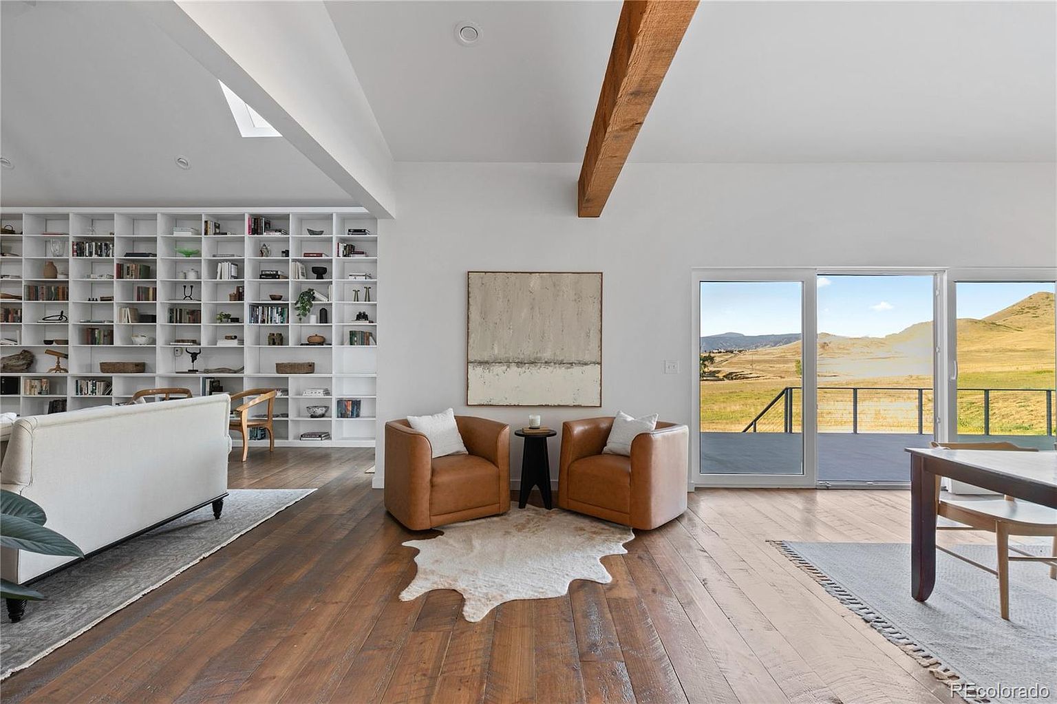 This is a bright and airy living room featuring hardwood floors, a large built-in bookshelf, and a sliding glass door that opens to a scenic view. The room is furnished with two leather armchairs, a small side table, and a modern sofa, creating a cozy and inviting atmosphere. A wooden beam adds a rustic touch to the modern design.