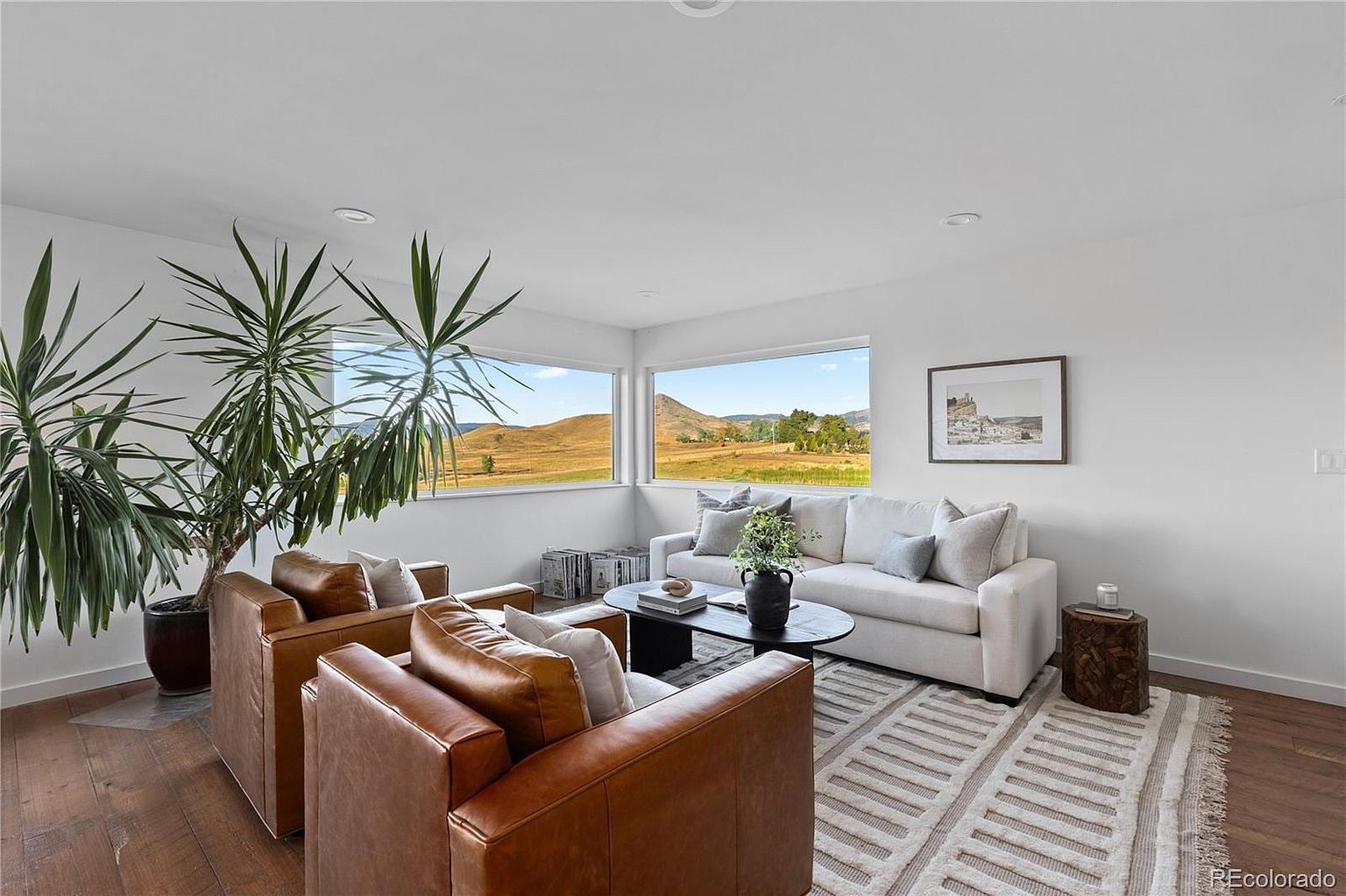 This is a bright and airy living room featuring two brown leather armchairs, a white sofa, and a black round coffee table. Large windows offer a scenic view of rolling hills and open fields, enhancing the room's connection to nature. The space is decorated in a minimalist style with a neutral color palette, creating a serene and inviting atmosphere.