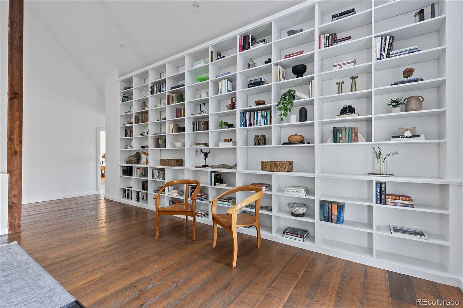 This interior shot showcases a living room with a large, built-in white bookshelf filled with books and decorative items. Two wooden chairs are positioned in front of the bookshelf, adding a touch of warmth to the space. The hardwood floors and natural light create an inviting and cozy atmosphere.