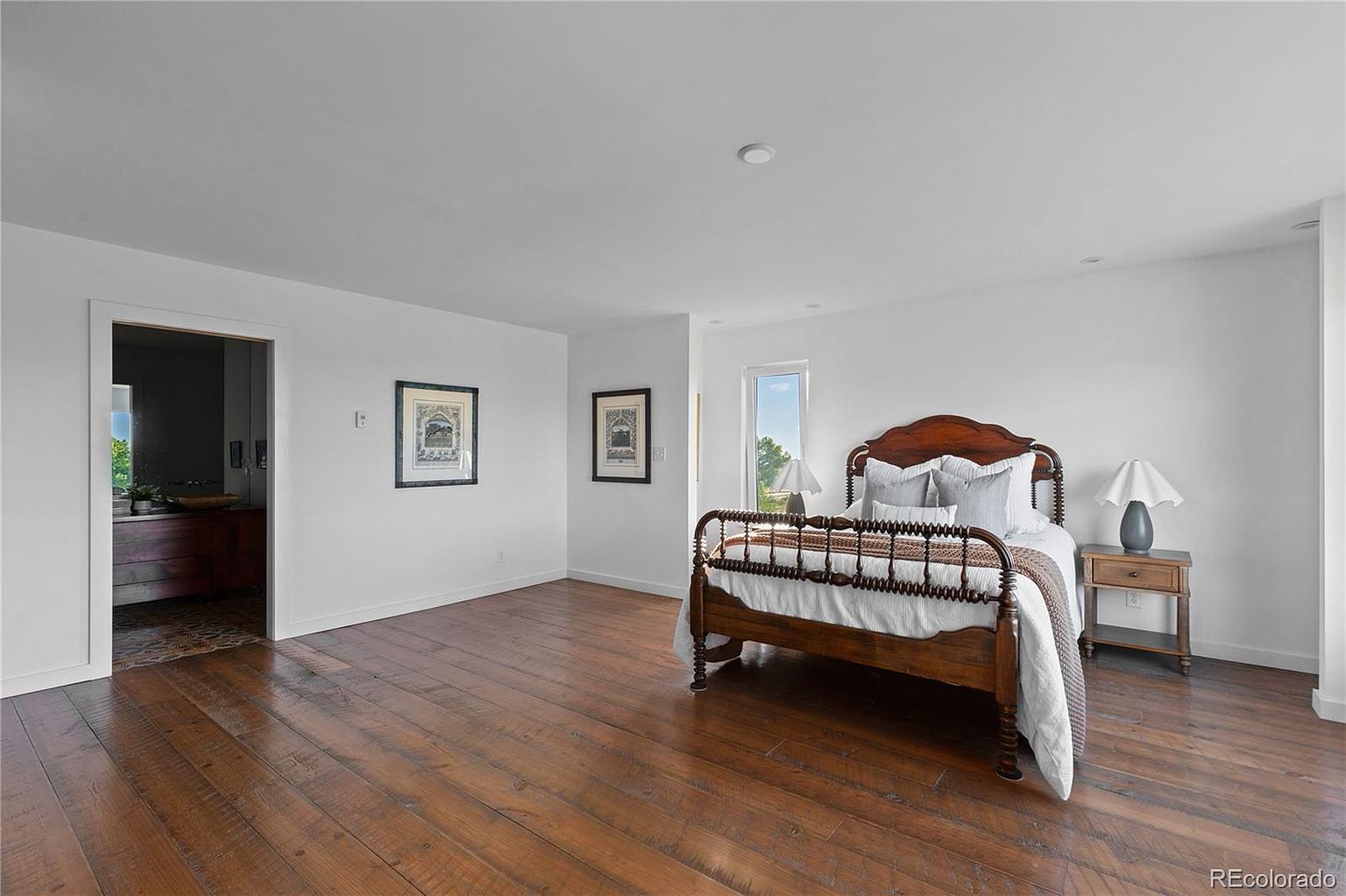 This is a primary bedroom featuring a wooden bed frame with white linens and decorative pillows. The room has hardwood floors, white walls, and two framed pictures. A small nightstand with a lamp sits beside the bed, and a doorway leads to another room, creating a spacious and inviting atmosphere.