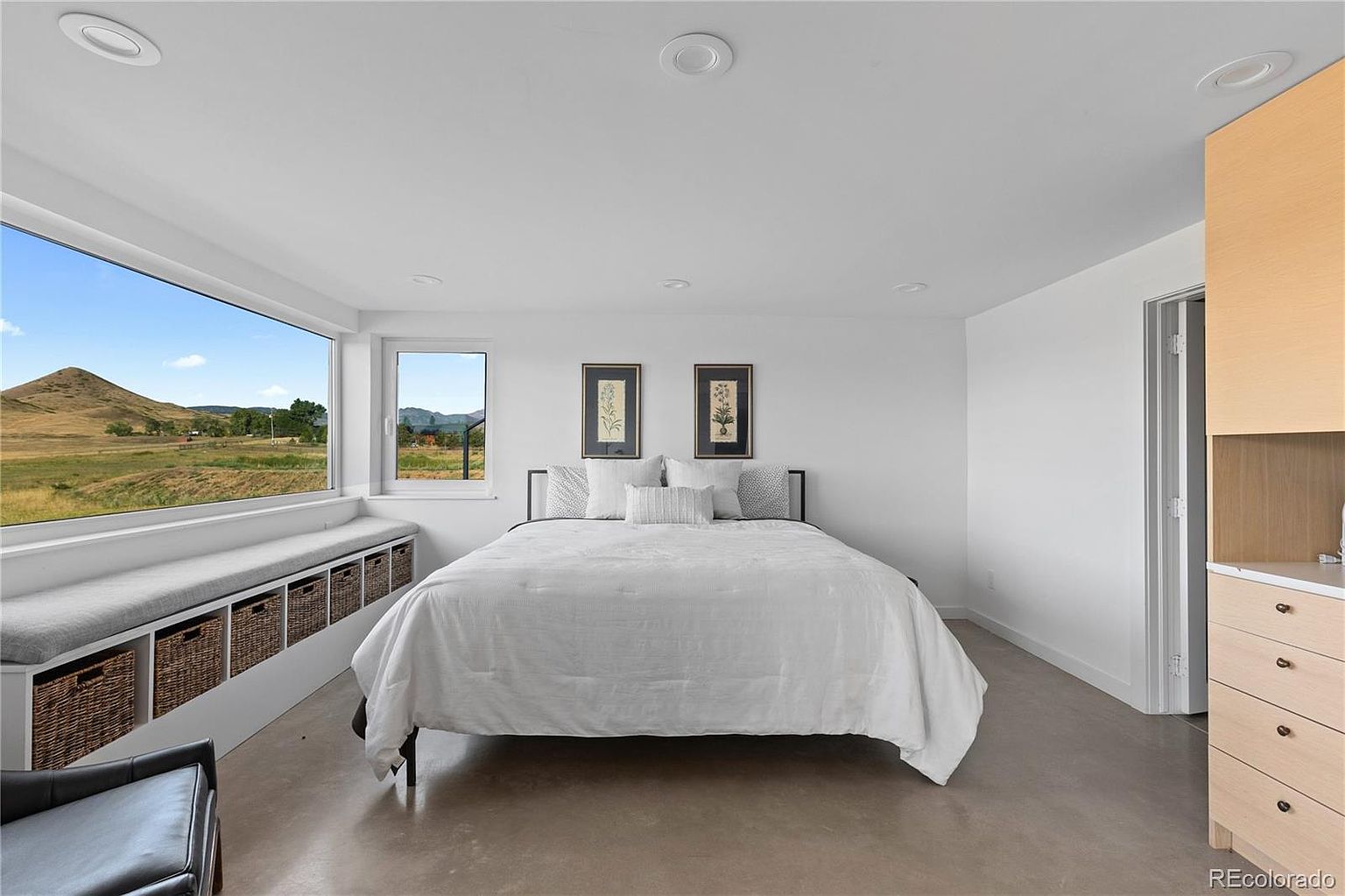 This is a bright and airy primary bedroom featuring a large window with a scenic view. The room is minimally decorated with a bed, two framed botanical prints, and a built-in window seat with storage. The polished concrete floors and white walls contribute to a modern and clean aesthetic.