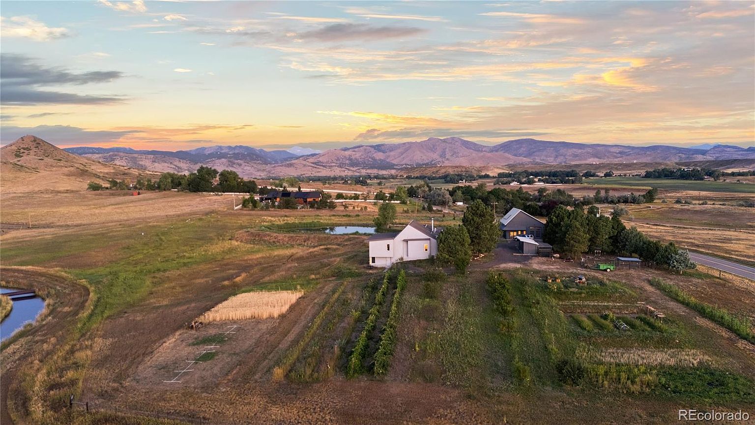 This aerial view showcases a sprawling property featuring modern farmhouses, cultivated fields, and a serene pond, all set against a backdrop of rolling hills and a vibrant sunset. The landscape includes a mix of agricultural plots, mature trees, and a well-maintained yard, creating a picturesque and tranquil estate. The perspective is from above, looking down at a slight angle, providing a comprehensive view of the property's layout and surrounding natural beauty.