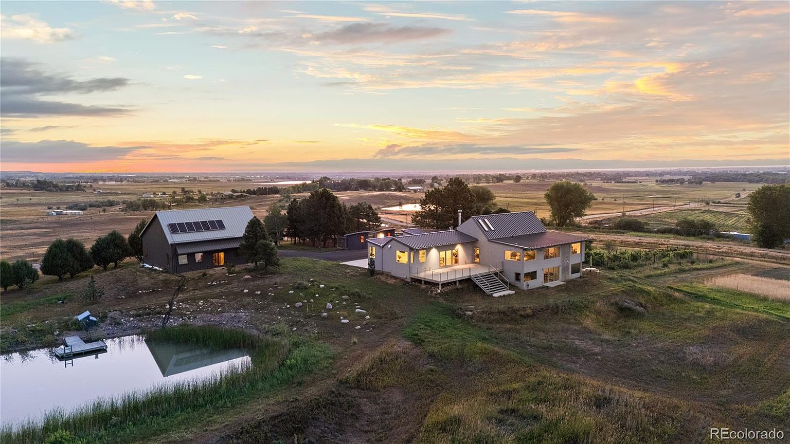 This aerial view showcases a stunning property featuring a modern home with a metal roof, complemented by a separate barn-like structure. The landscape includes a serene pond with a dock, rolling grassy hills, and distant fields under a picturesque sunset sky. The overall impression is one of peaceful, rural luxury.