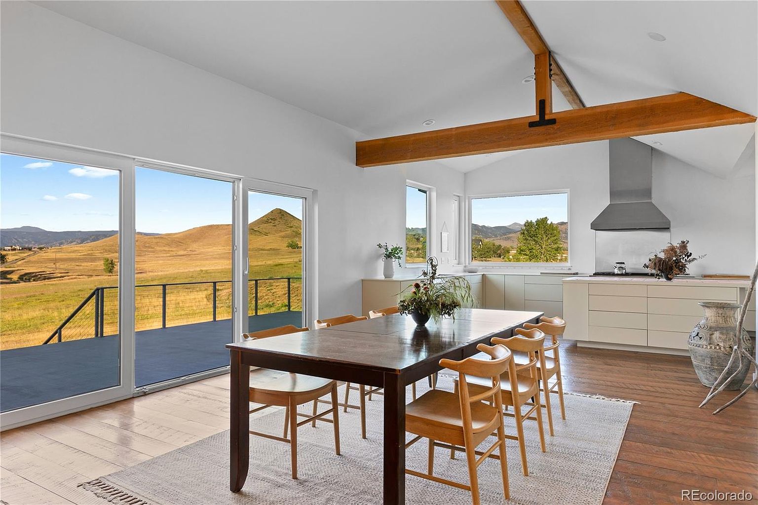 This interior shot showcases a dining room with a dark wood table and light wood chairs set on a neutral rug. Large sliding glass doors offer a stunning view of the landscape, while a wooden beam adds architectural interest to the white-walled space. The room seamlessly blends indoor comfort with outdoor scenery.