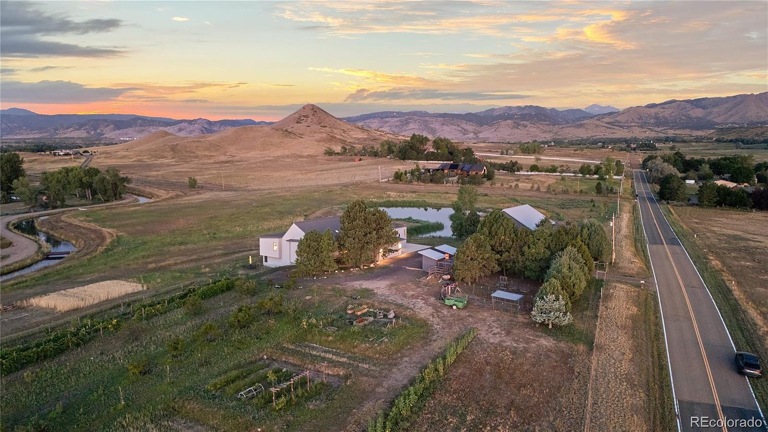 This aerial view showcases a sprawling property featuring a modern farmhouse-style home with a pond, outbuildings, and cultivated gardens. The landscape includes rolling hills and distant mountains under a vibrant sunset sky. A nearby road adds to the property's accessibility, while the overall impression is one of serene rural living.