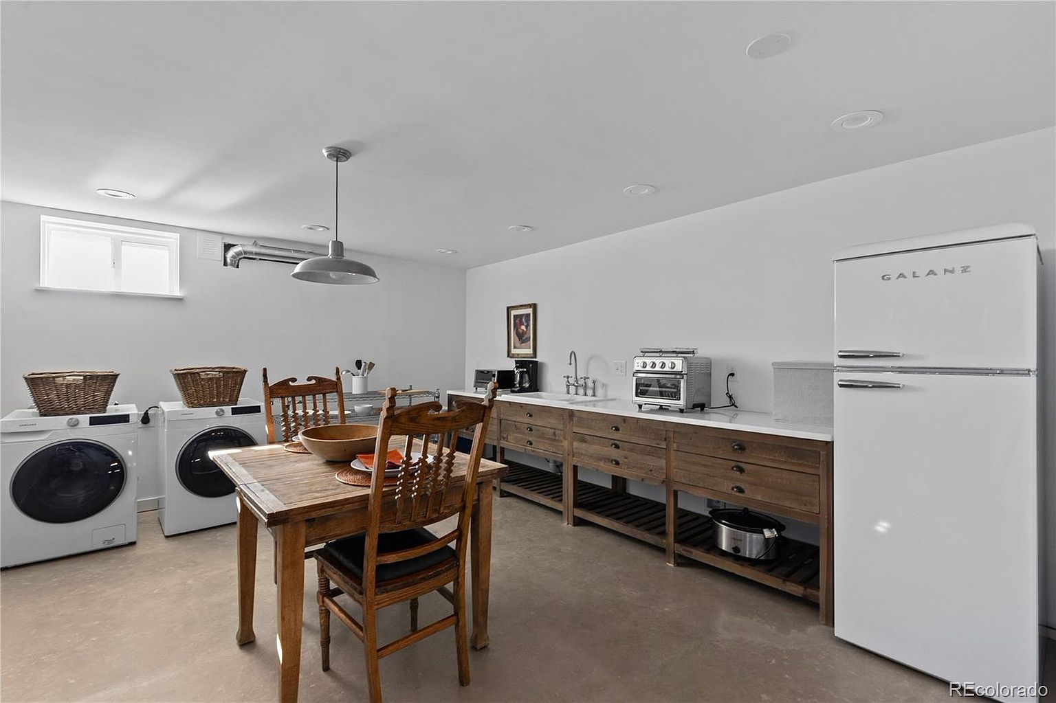This interior shot showcases a unique kitchen area that blends functionality with a rustic aesthetic. The space features a wooden dining table with chairs, a long wooden countertop with a sink, toaster oven, and other appliances, and a vintage-style refrigerator. Adjacent to the kitchen area are modern washing machines, creating an unexpected yet practical combination of spaces.