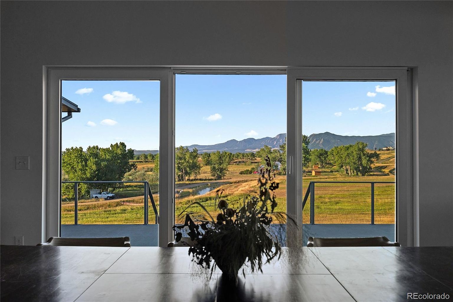 The image showcases a dining room with a large wooden table as the focal point, adorned with a floral arrangement. The room features a wall of windows offering a scenic view of a landscape with fields, trees, and distant mountains under a blue sky. The overall impression is one of tranquility and connection to nature.